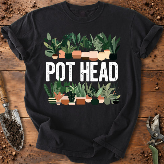 A black short-sleeve graphic T-shirt laid flat on a rustic wooden surface with gardening props. The shirt features a colorful illustration of potted plants above and below large white text reading “POT HEAD.