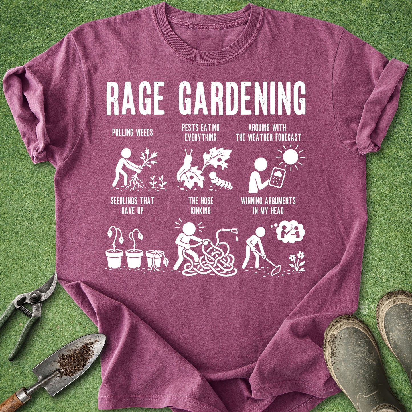 A maroon short-sleeve t-shirt with a white 'RAGE GARDENING' graphic print, laid flat on a green grass background with gardening tools like pruning shears, a trowel, and boots.