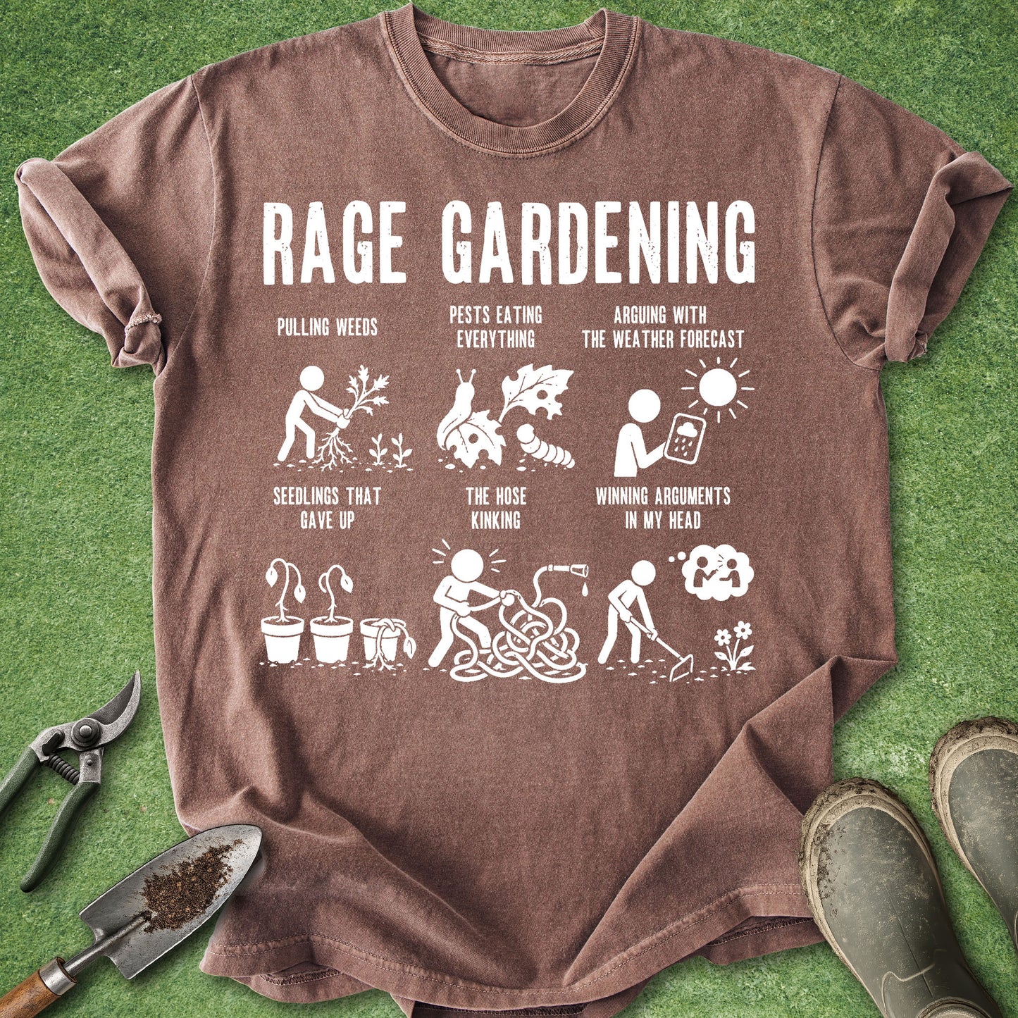 A brown short-sleeve graphic T-shirt laid flat on green grass-like background, with gardening-themed white text and illustrations reading 'RAGE GARDENING' and smaller slogan panels. Gardening props including pruning shears, a trowel, and boots are placed around the shirt.