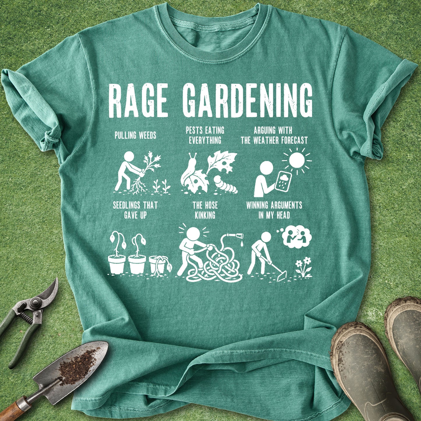A teal green graphic t-shirt laid flat on a green grass-like surface, surrounded by gardening props including pruning shears, a trowel, and work boots. The shirt features a white graphic with the text 'RAGE GARDENING' and several humorous gardening illustrations and captions.