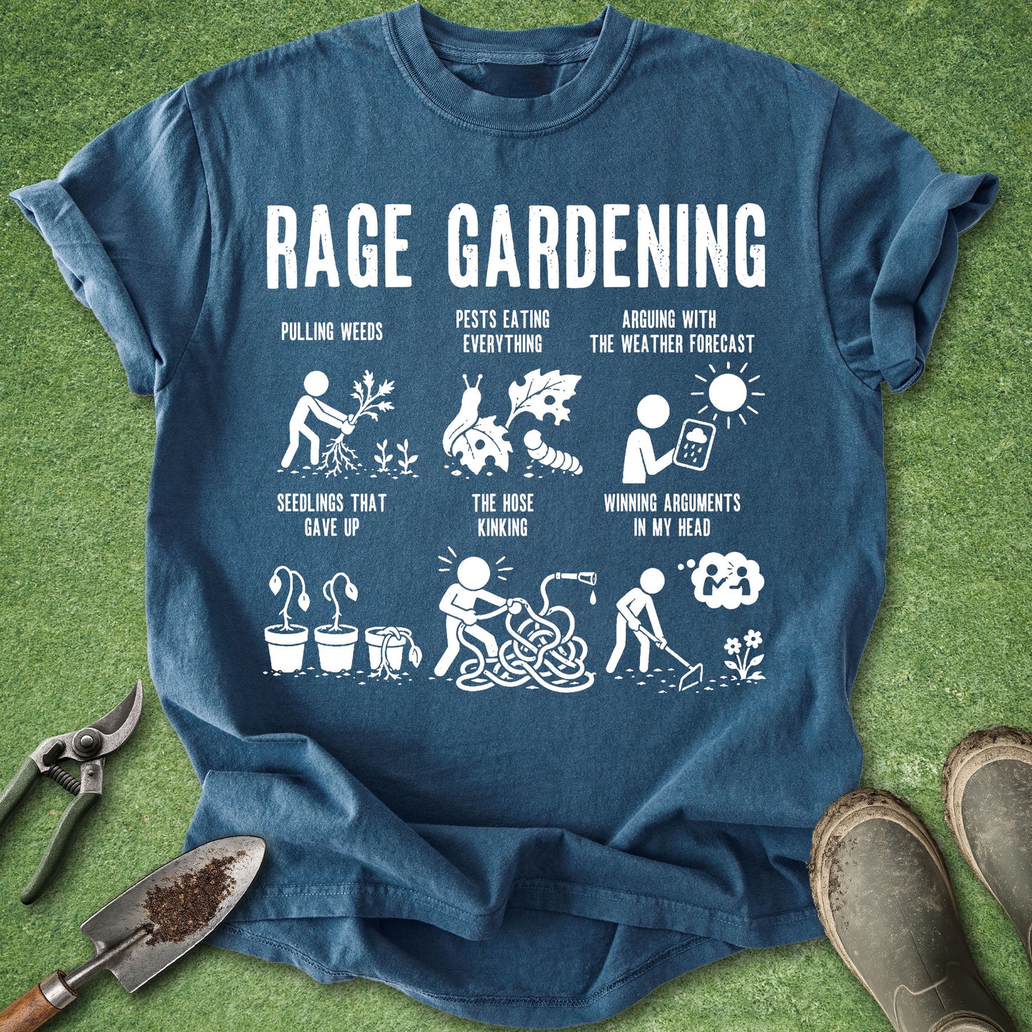 A blue graphic t-shirt laid flat on green grass with gardening props around it, including pruning shears, a trowel, and work boots. The shirt features a white 'RAGE GARDENING' graphic with humorous illustrations and text related to gardening challenges.