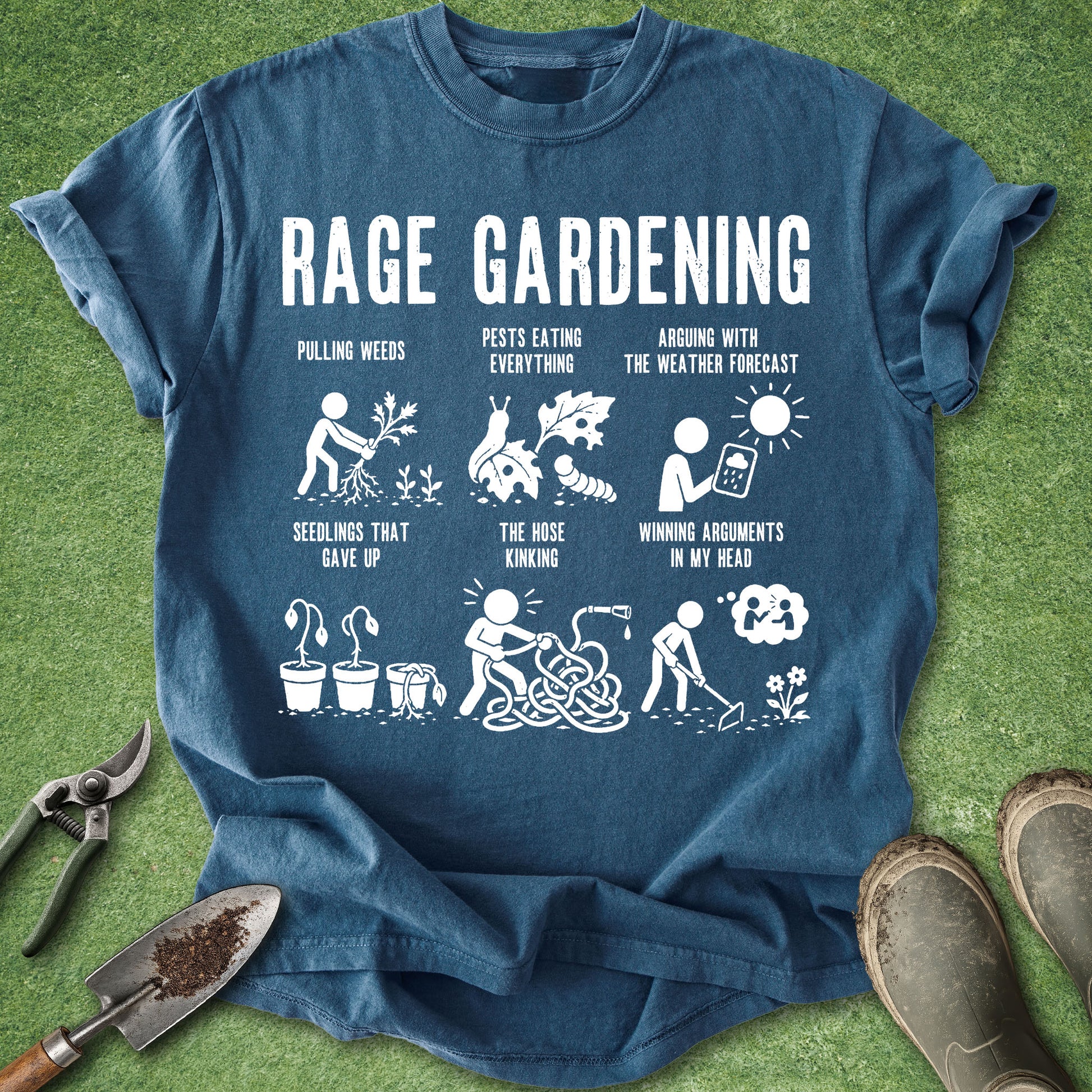 A blue graphic t-shirt laid flat on green grass with gardening props around it, including pruning shears, a trowel, and work boots. The shirt features a white 'RAGE GARDENING' graphic with humorous illustrations and text related to gardening challenges.