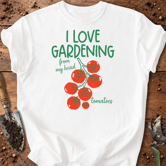 A white short-sleeve graphic T-shirt laid flat on a rustic wooden surface with gardening props. The shirt features green text reading 'I LOVE GARDENING from my head tomatoes' above an illustration of red tomatoes on a vine.