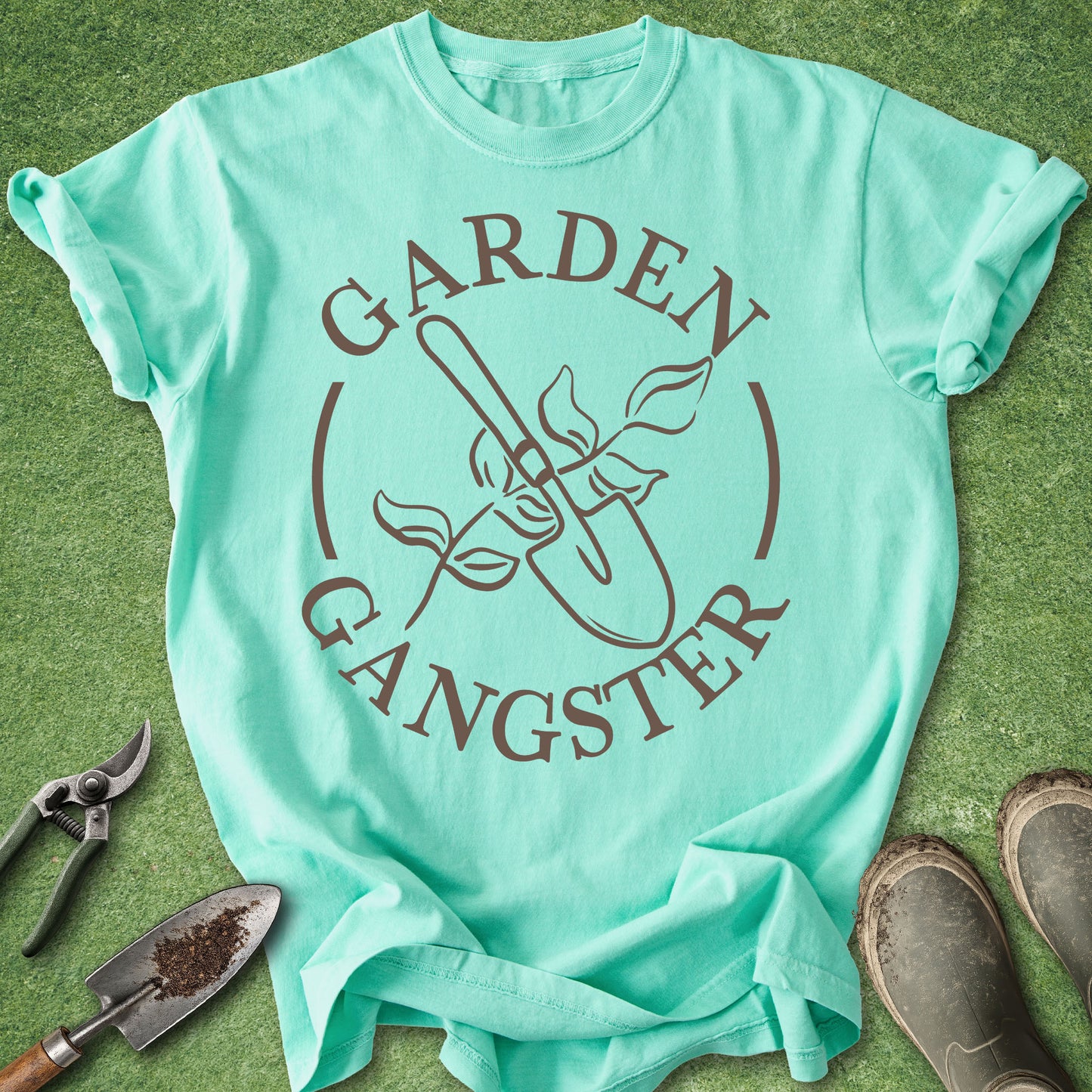 Mint green t-shirt with 'Garden Gangster' design on a grass background with gardening tools.