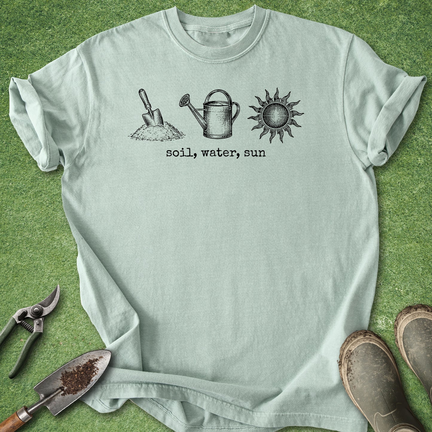 A light sage green t-shirt with a black graphic of a shovel, watering can, and sun with the text 'soil, water, sun' printed on the chest. The shirt is laid flat on a green grass-like surface with gardening props (shears, trowel, boots) around it.