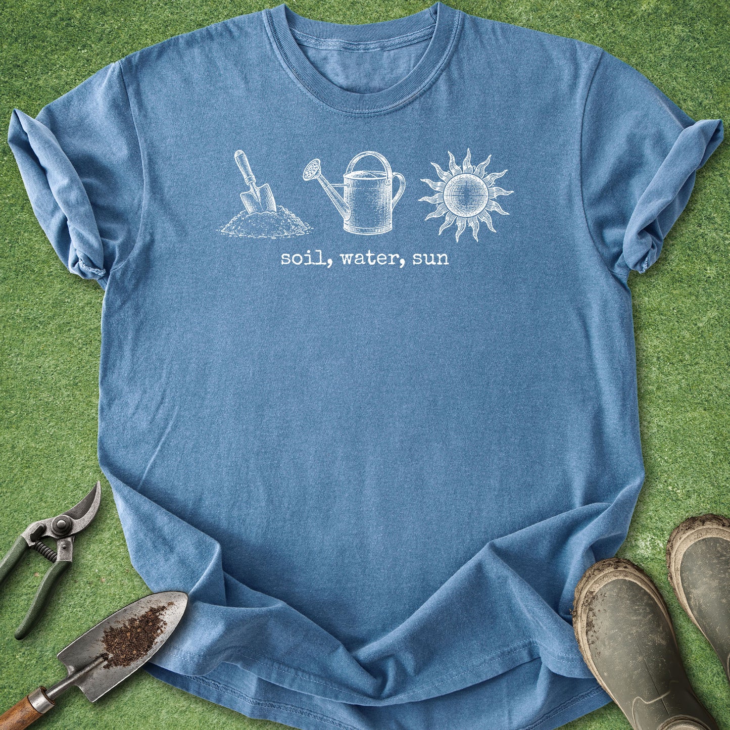 A blue short-sleeve t-shirt laid flat on green grass, featuring a white graphic of a shovel, watering can, and sun with the text 'soil, water, sun'. Gardening tools and muddy boots are placed around the shirt as styling props.