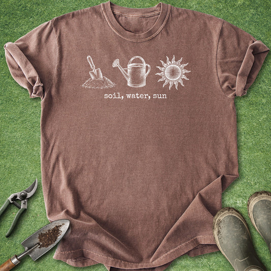 A heathered brown short-sleeve t-shirt laid flat on green grass, featuring a white line art graphic of a shovel, watering can, and sun with the text 'soil, water, sun' printed below. Gardening props including pruning shears, a trowel with soil, and muddy boots are arranged around the shirt.