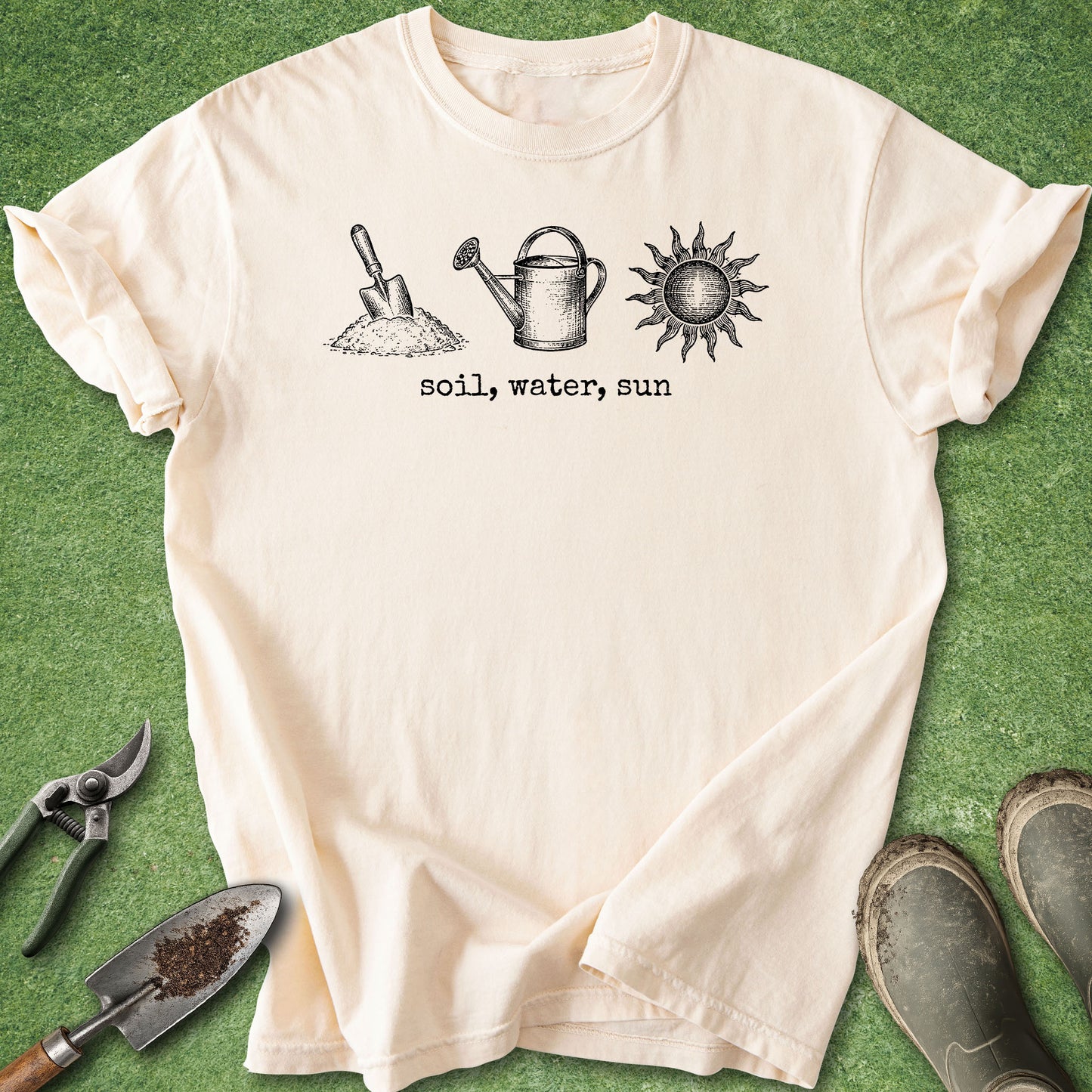 A cream-colored short-sleeve t-shirt with a black graphic of a shovel, watering can, and sun with the text 'soil, water, sun' underneath. The shirt is laid flat on a green grass-like surface, surrounded by gardening props including pruning shears, a trowel, and boots.