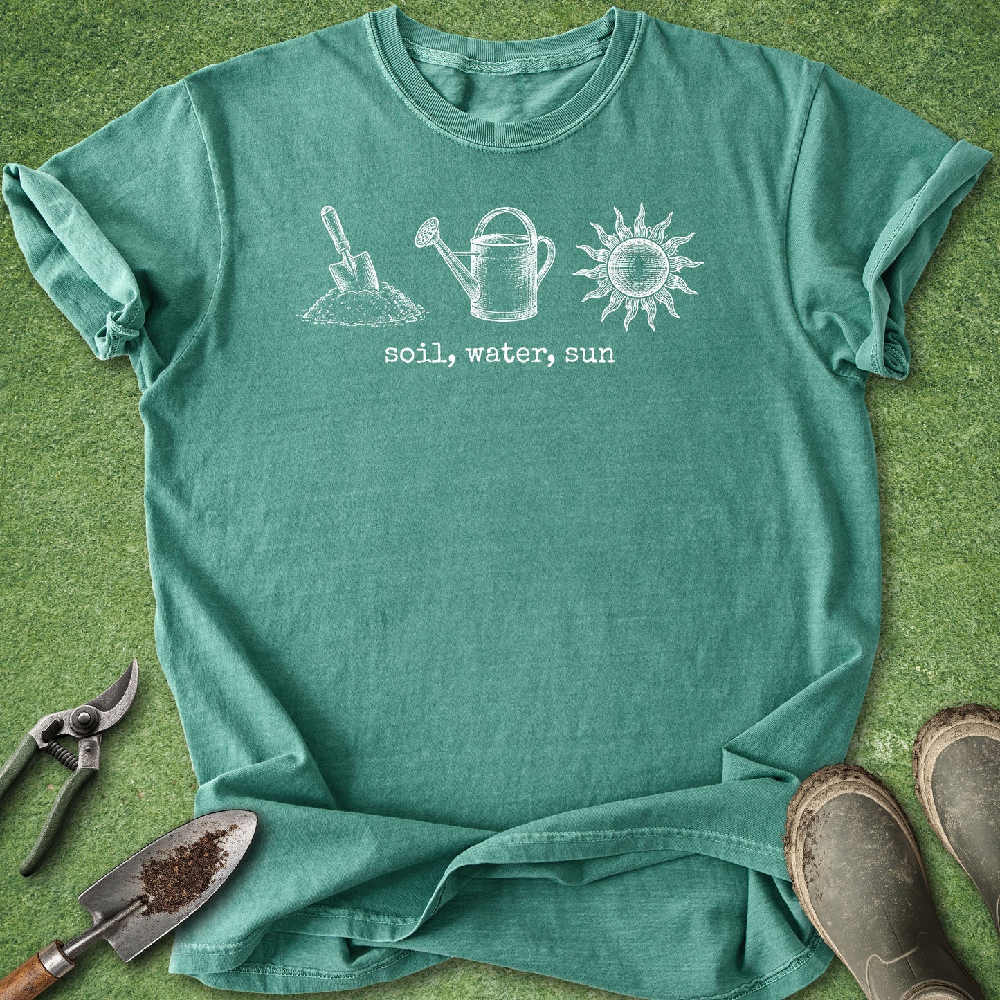 A teal green short-sleeve t-shirt with a white graphic of gardening tools and the text 'soil, water, sun' laid flat on a green grass-like background, surrounded by gardening props including pruning shears, a trowel, and boots.