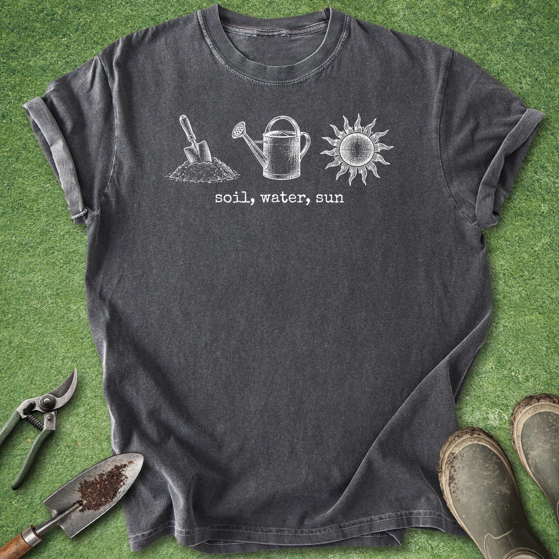 A dark charcoal gray t-shirt with a white graphic of a gardening shovel, watering can, and sun with the text 'soil, water, sun' printed on the chest. The shirt is laid flat on green grass with gardening props including pruning shears, a trowel, and boots.