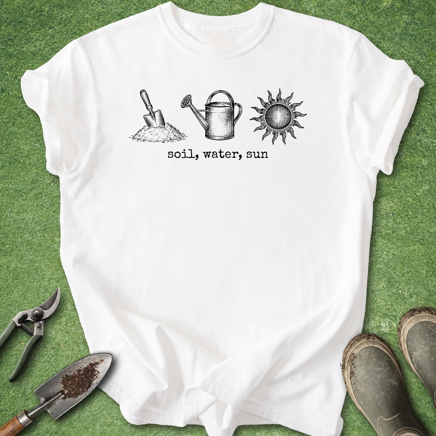 A white short-sleeve T-shirt laid flat on green grass, featuring a black line-art graphic of a shovel in soil, a watering can, and a sun with the text 'soil, water, sun' underneath. Gardening props including pruning shears, a trowel, and boots are placed around the shirt.