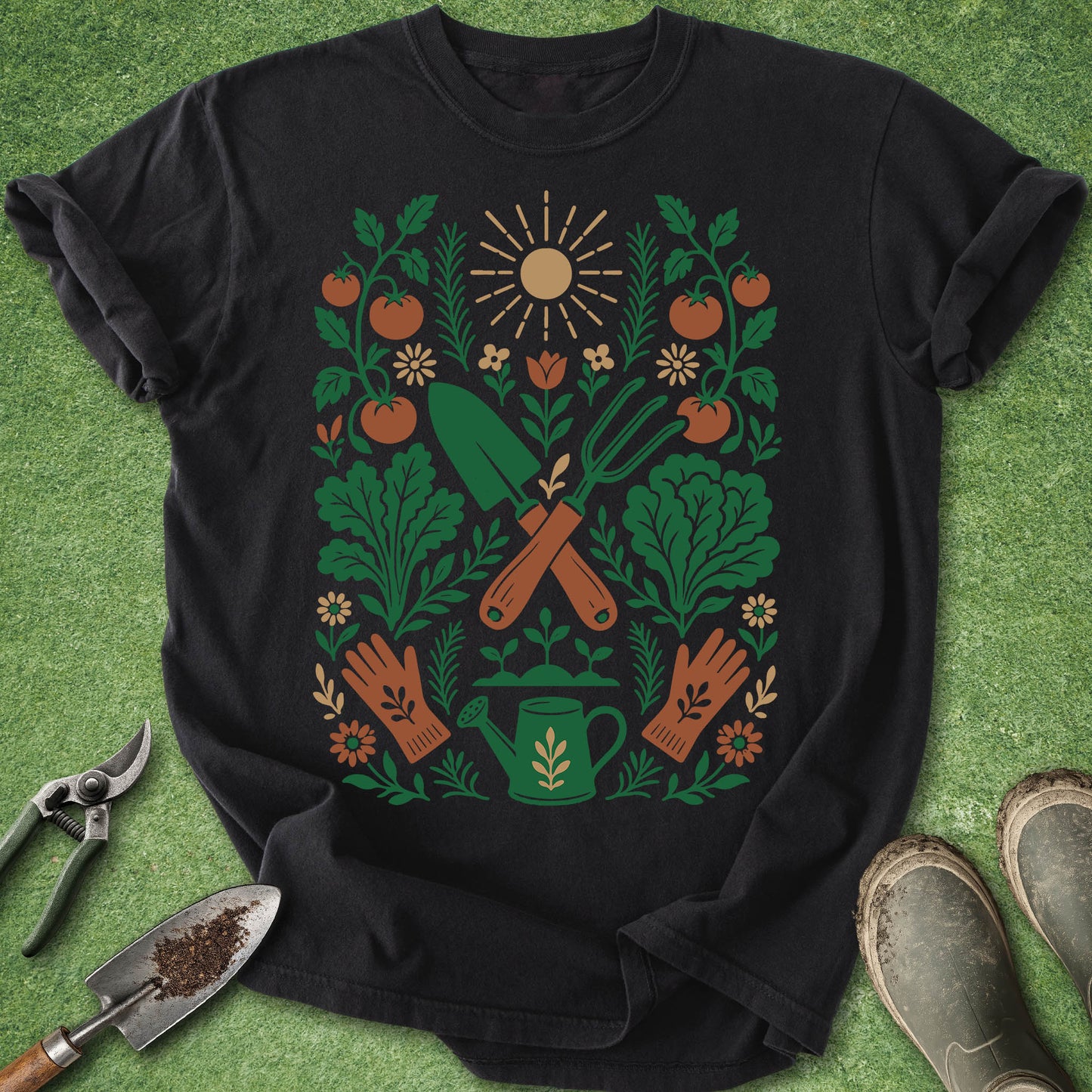 A black short-sleeve graphic T-shirt laid flat on a green textured surface, surrounded by gardening props including pruning shears, a trowel, and work boots. The shirt features a large front print with green leaves, orange tomatoes and flowers, brown crossed tools, and a green watering can.