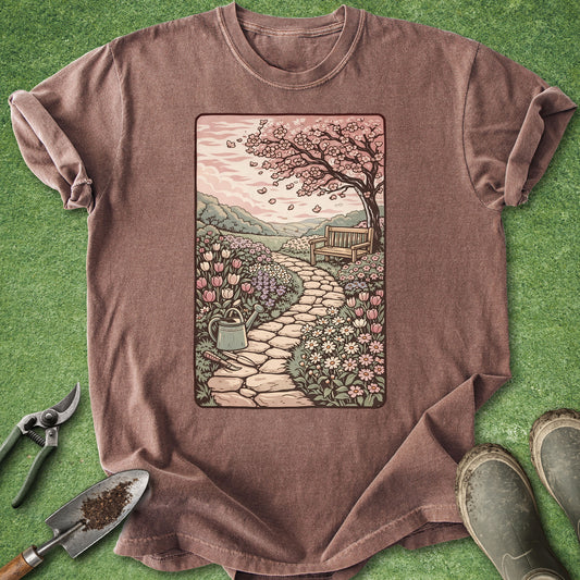 Vintage-style garden illustration t-shirt featuring a blooming flower path, wooden bench, and cherry blossom tree in soft pastel colors on a mauve shirt