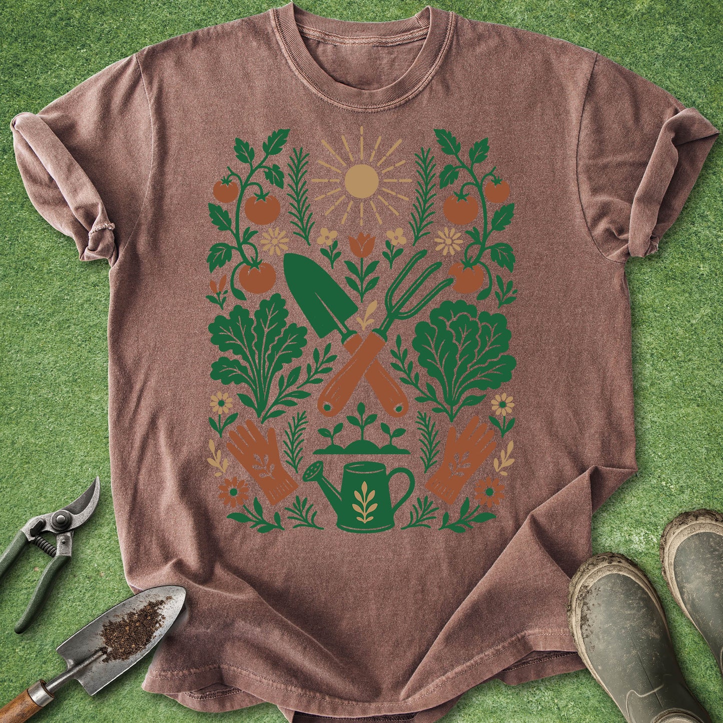 A heathered brown short-sleeve t-shirt laid flat on green grass, featuring a colorful gardening graphic with a sun, tomatoes, leafy plants, a watering can, and tools. A small gardening trowel and pruning shears are placed to the left, and muddy boots are at the bottom right.