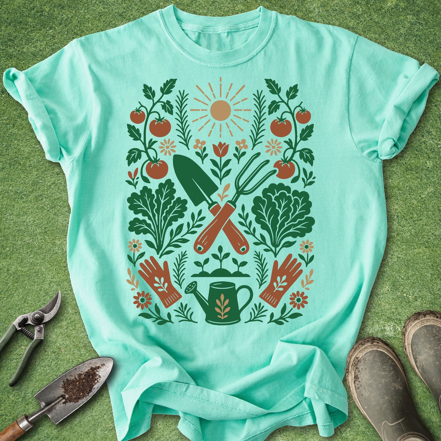 A mint green short-sleeve t-shirt with a colorful gardening graphic print, laid flat on a green grass-like surface with gardening props including pruning shears, a trowel, and boots.