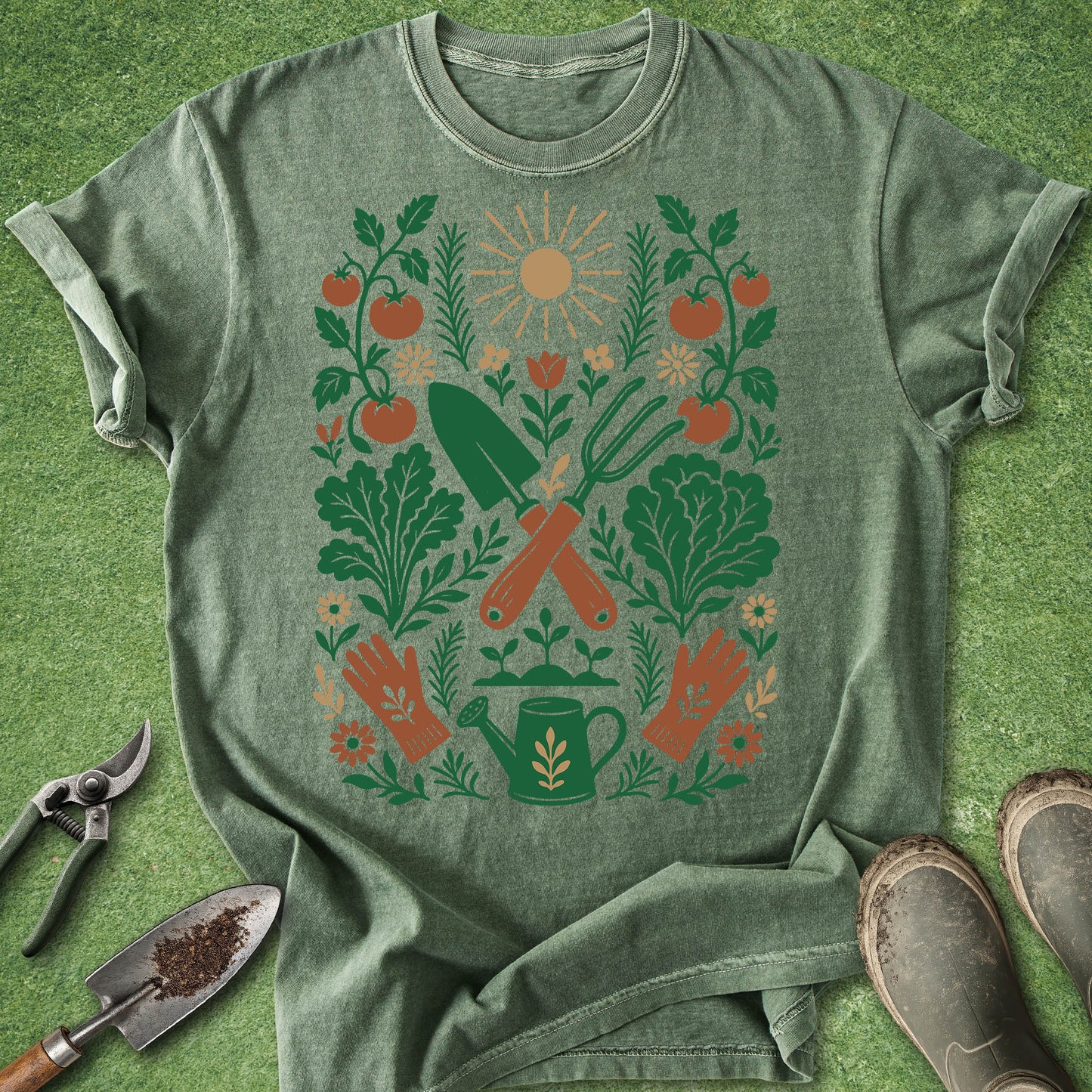 A heathered sage green t-shirt laid flat on a green grass-like surface, featuring a colorful gardening graphic with a sun, tomatoes, leafy greens, and tools. A gardening shears, trowel, and boots are styled around the shirt.