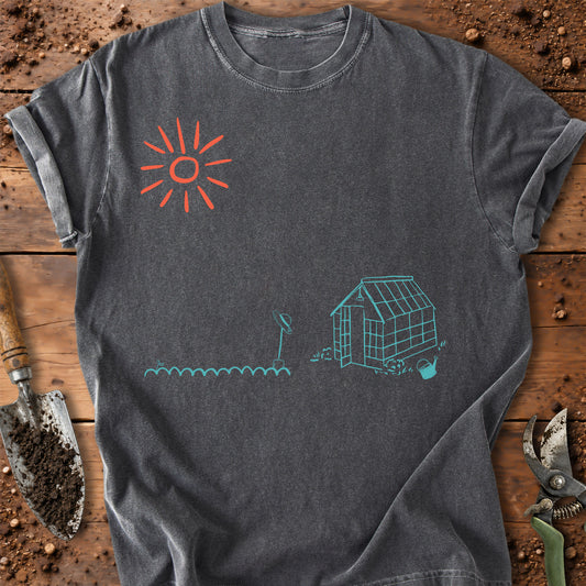 A dark gray t-shirt laid flat on a rustic wooden surface with gardening props like a trowel and pruning shears. The shirt features a simple graphic print of a red sun and a teal greenhouse and plant design.