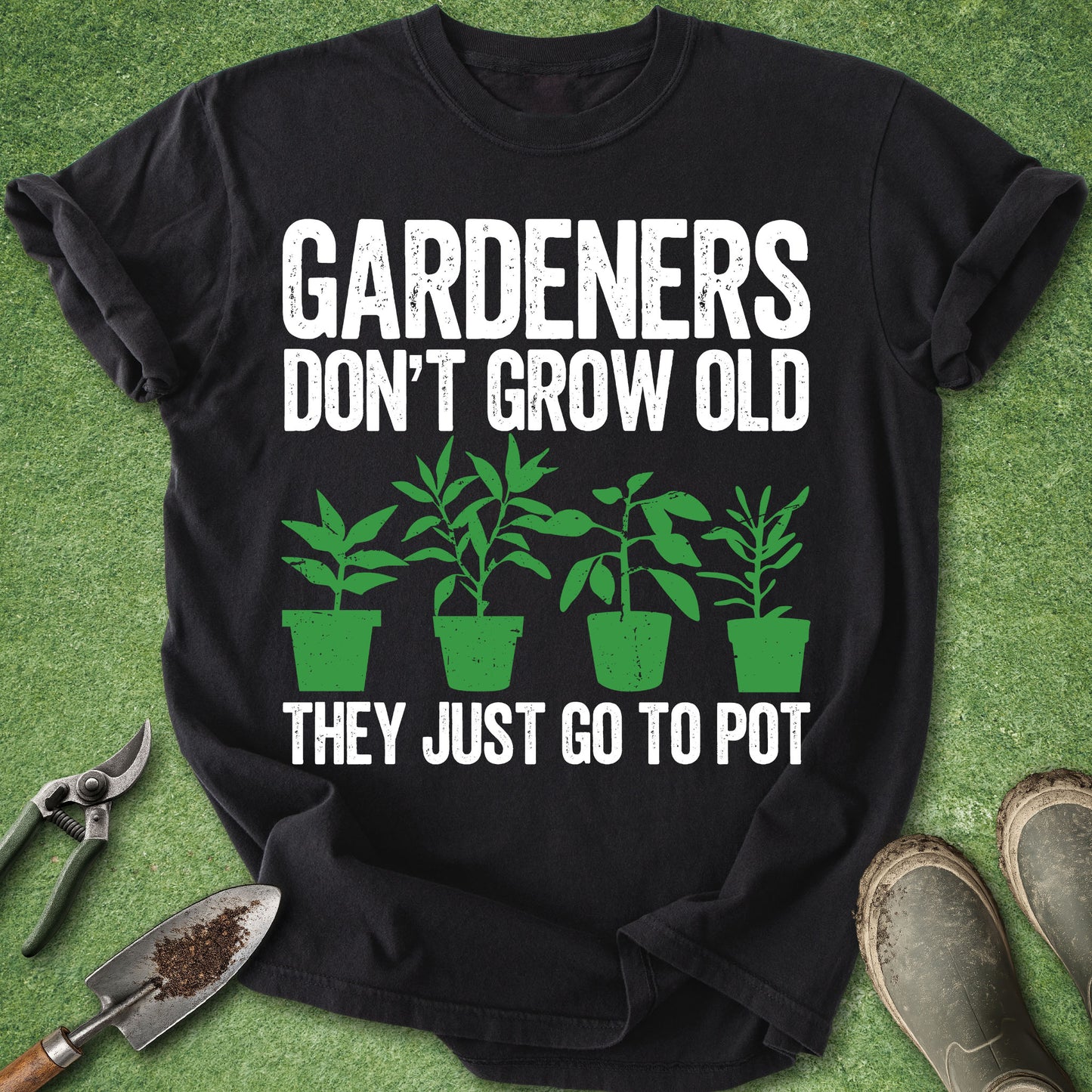 A black short-sleeve graphic T-shirt laid flat on green grass-like background, with white text reading “GARDENERS DON’T GROW OLD THEY JUST GO TO POT” and green plant illustrations. Gardening props including pruning shears, a trowel, and boots are placed around the shirt.