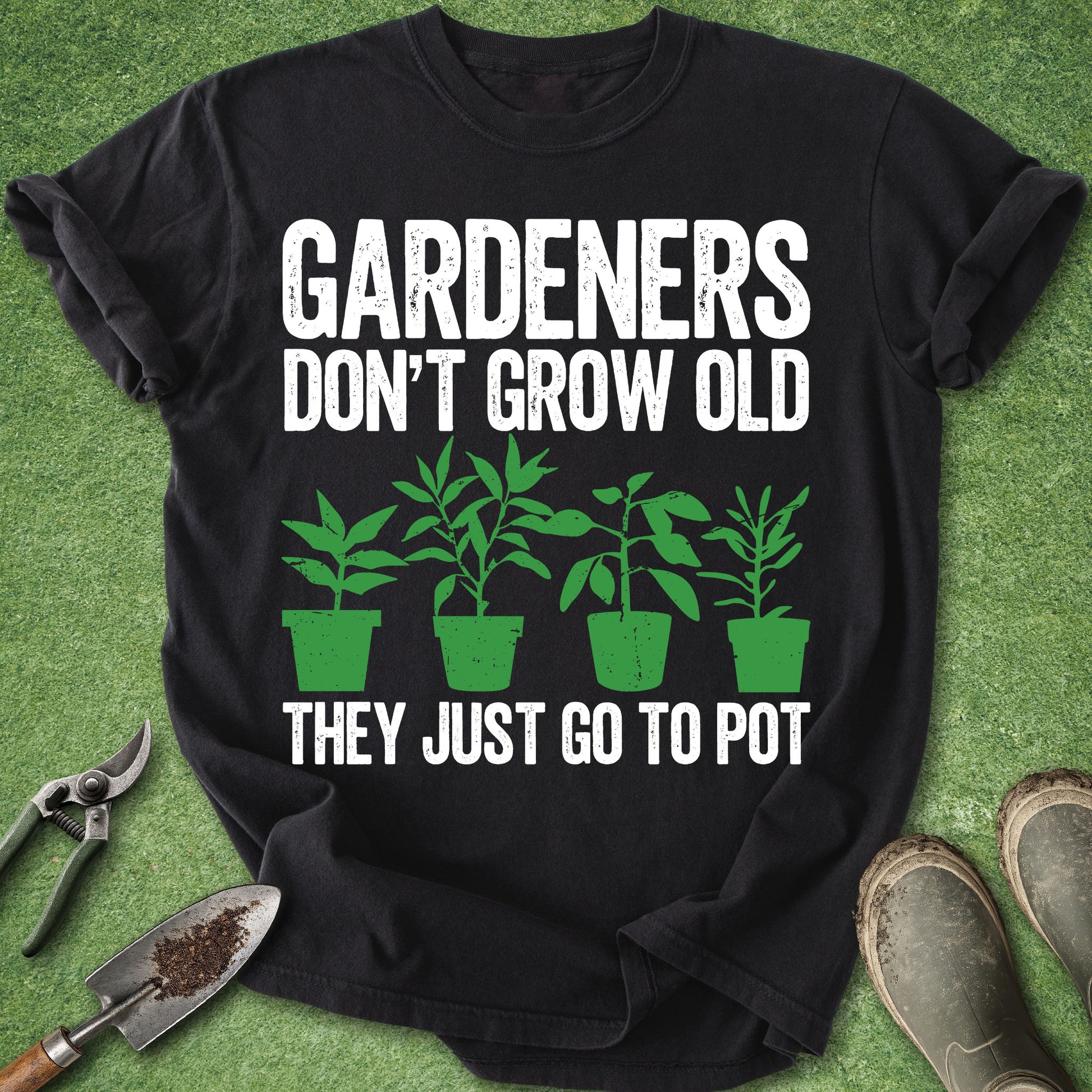 A black short-sleeve graphic T-shirt laid flat on green grass-like background, with white text reading “GARDENERS DON’T GROW OLD THEY JUST GO TO POT” and green plant illustrations. Gardening props including pruning shears, a trowel, and boots are placed around the shirt.