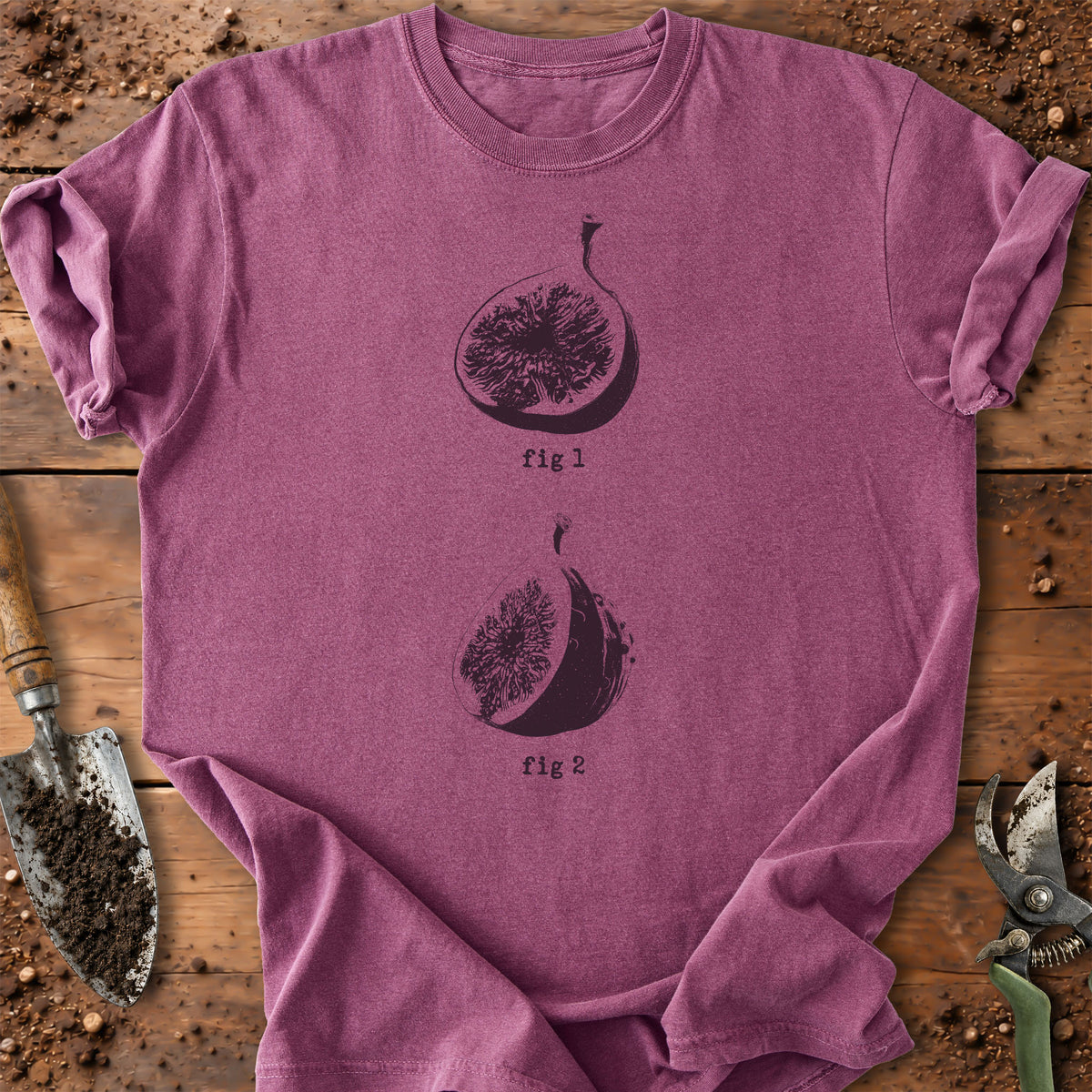 A heathered mauve-purple short-sleeve t-shirt with a black graphic of two figs labeled 'fig 1' and 'fig 2', laid flat on a rustic wooden surface with gardening tools nearby.