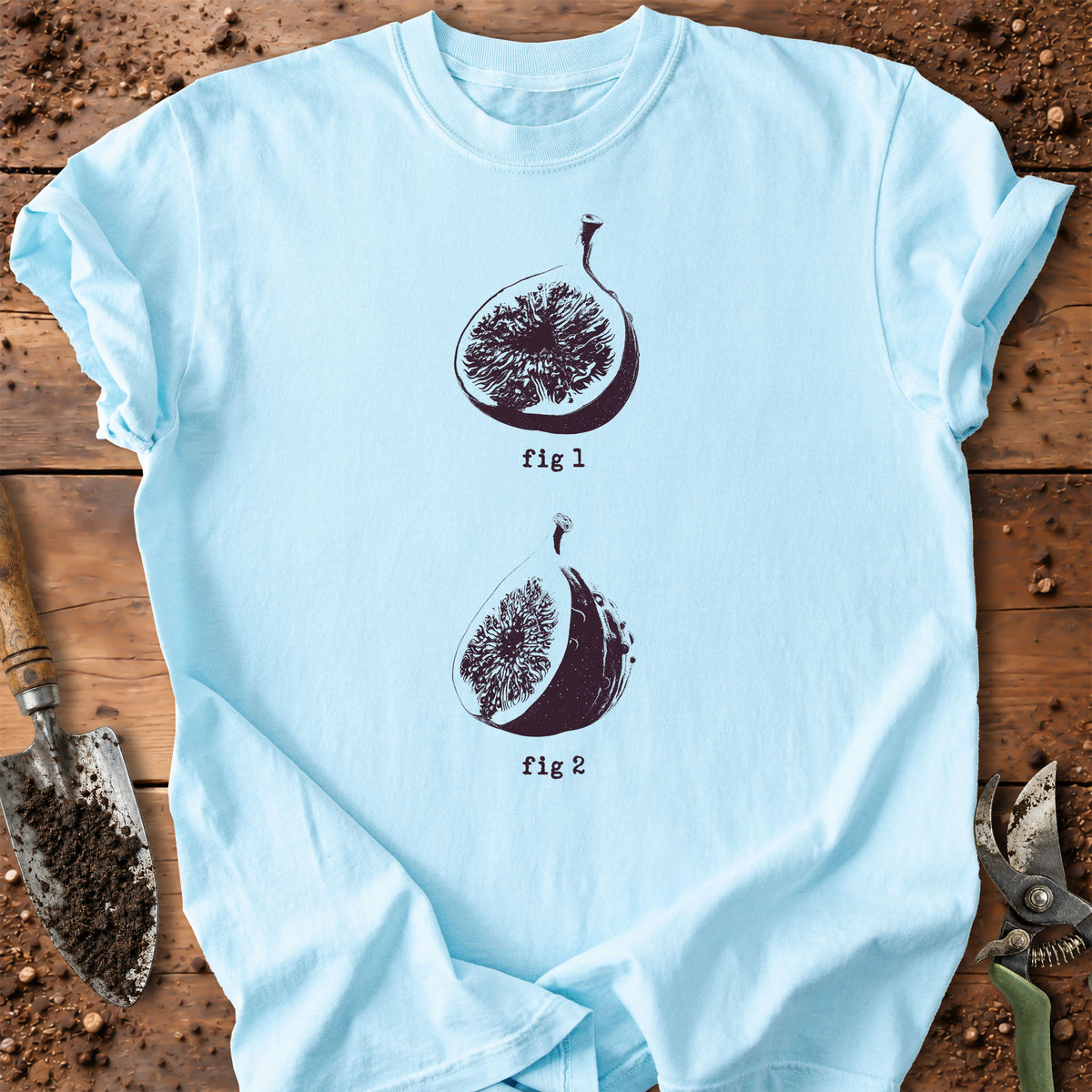 A light blue short-sleeve t-shirt laid flat on a rustic wooden surface with gardening props. The shirt features a dark brown graphic of two fig halves labeled 'fig 1' and 'fig 2'.