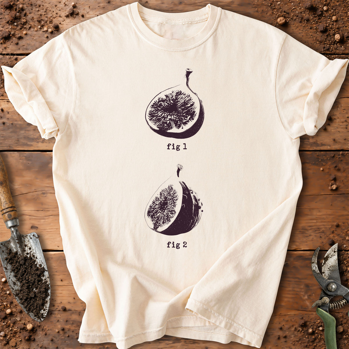 An off-white short-sleeve t-shirt with a dark purple graphic of two figs labeled 'fig 1' and 'fig 2', laid flat on a rustic wooden surface with gardening tools.