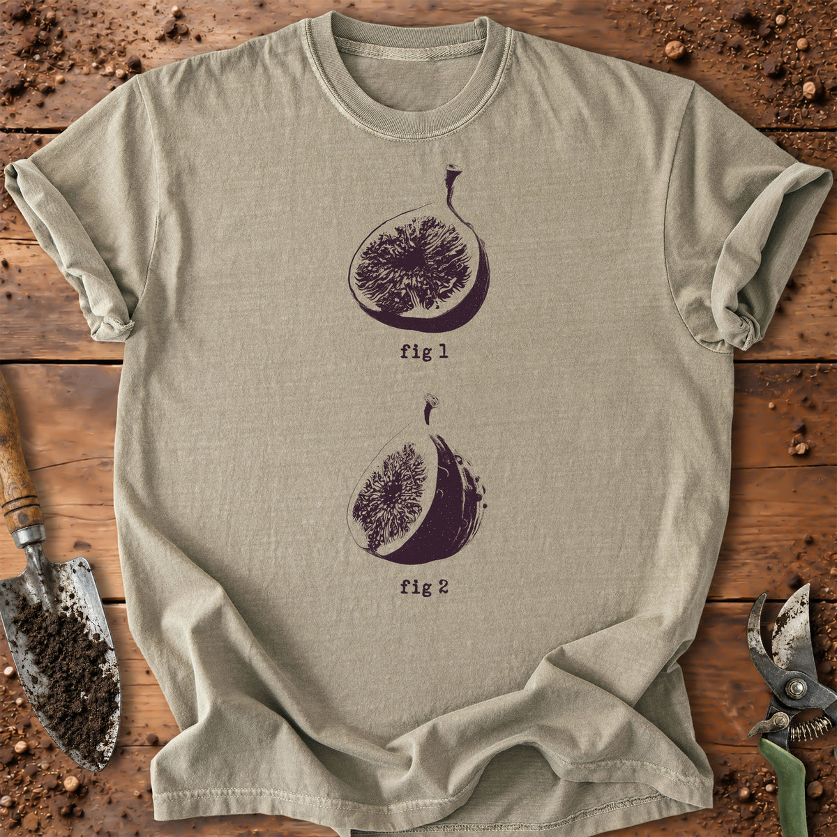 A khaki lavender crewneck t-shirt with a dark purple graphic of two figs labeled 'fig 1' and 'fig 2', laid flat on a rustic wooden surface with gardening tools.