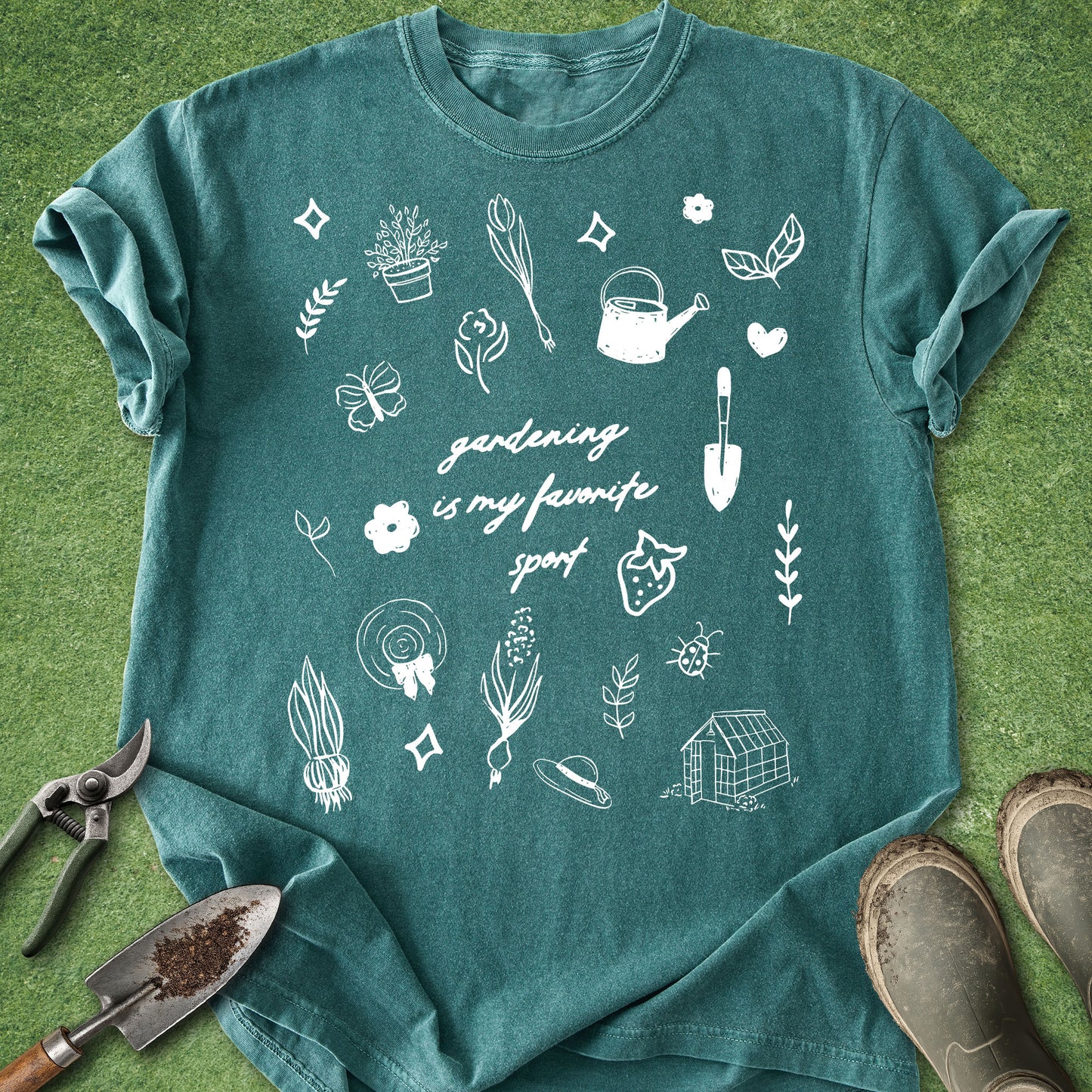 A teal short-sleeve t-shirt with white gardening-themed doodles and the text 'gardening is my favourite sport' laid flat on green grass, styled with gardening tools and boots.