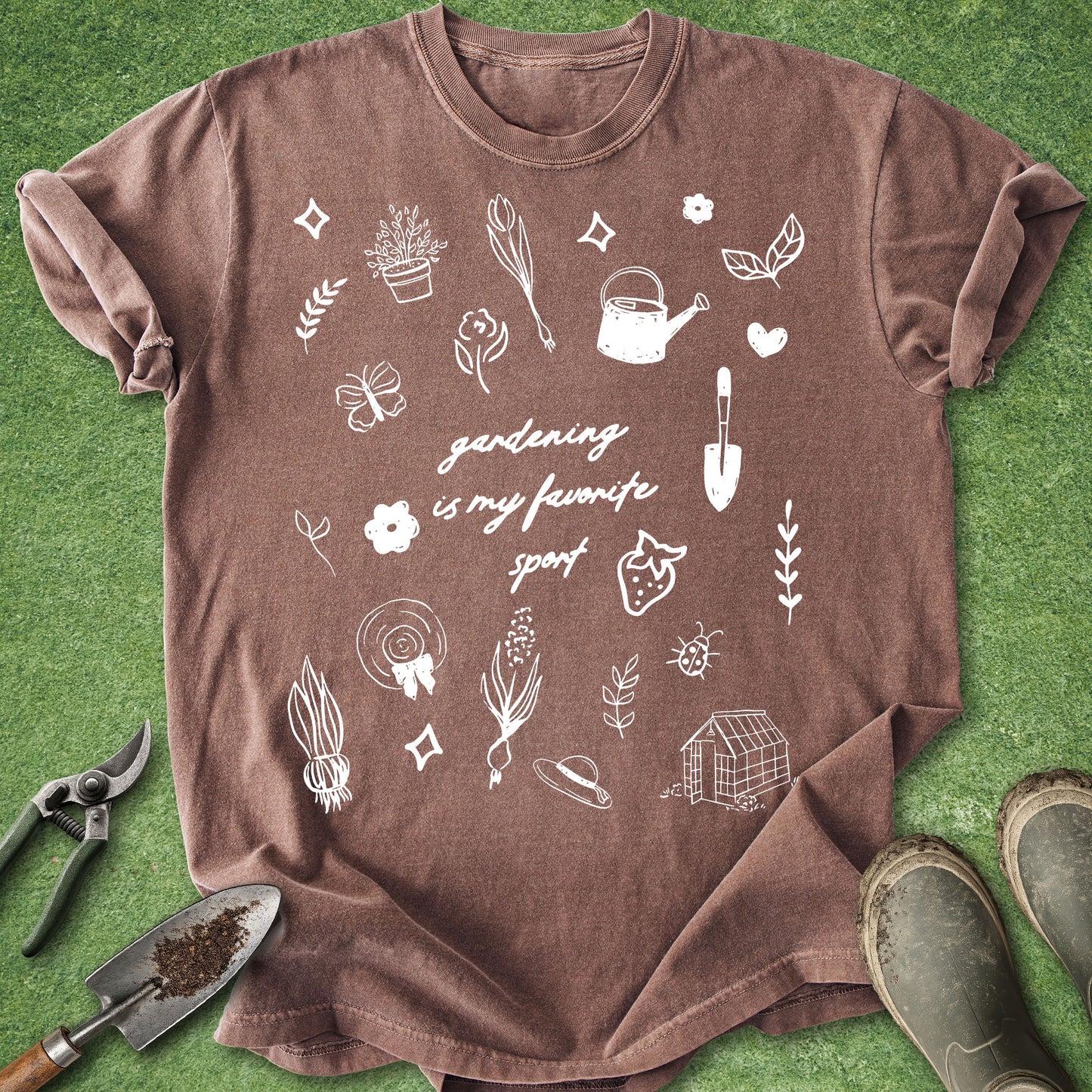 A brown short-sleeve t-shirt with white gardening-themed illustrations and the text 'gardening is my fav sport' laid flat on green grass, surrounded by gardening props including pruning shears, a trowel, and boots.