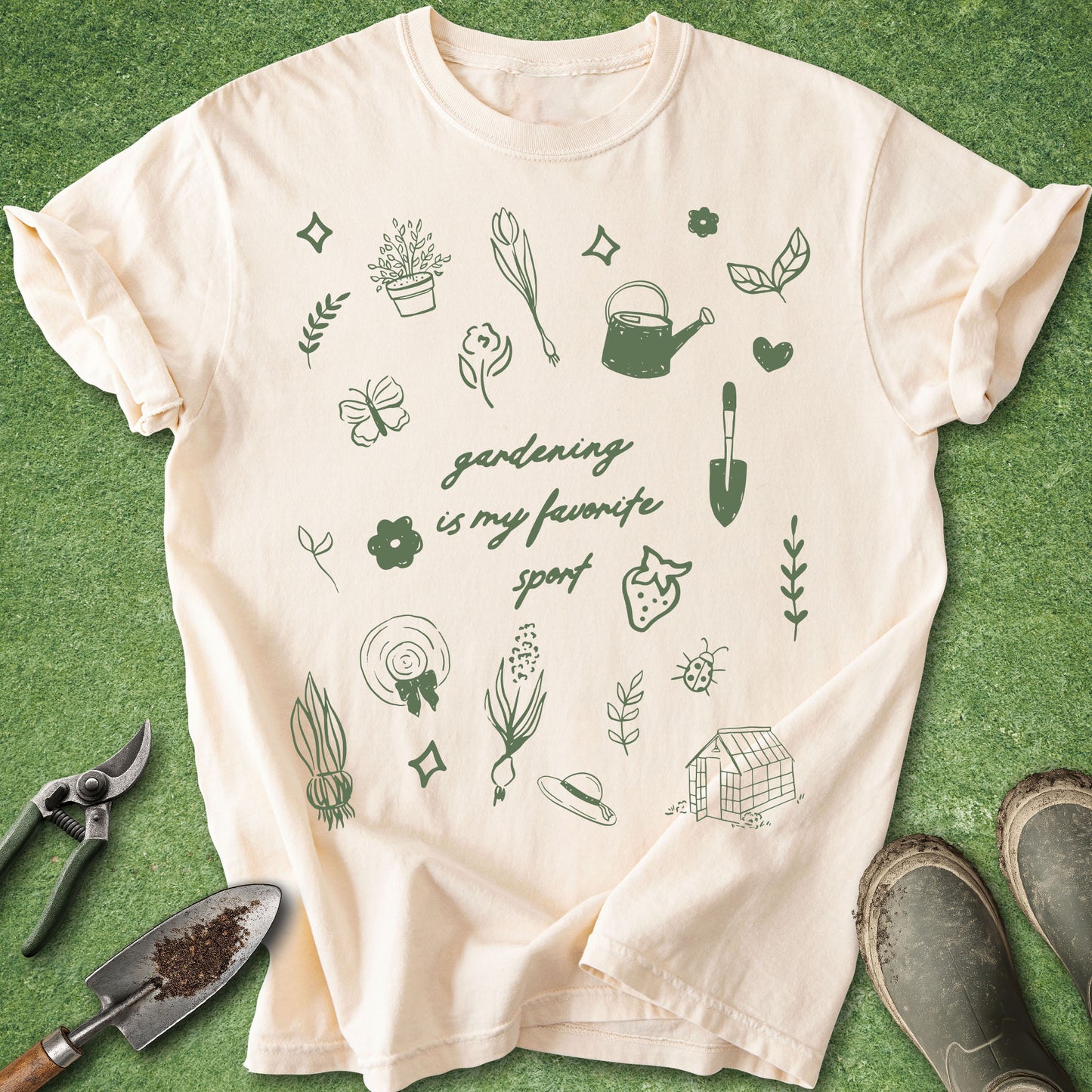 A cream-colored short-sleeve t-shirt with a green graphic print of gardening-themed doodles and the text 'gardening is my favorite sport', laid flat on a green grass-like surface with gardening props and boots nearby.
