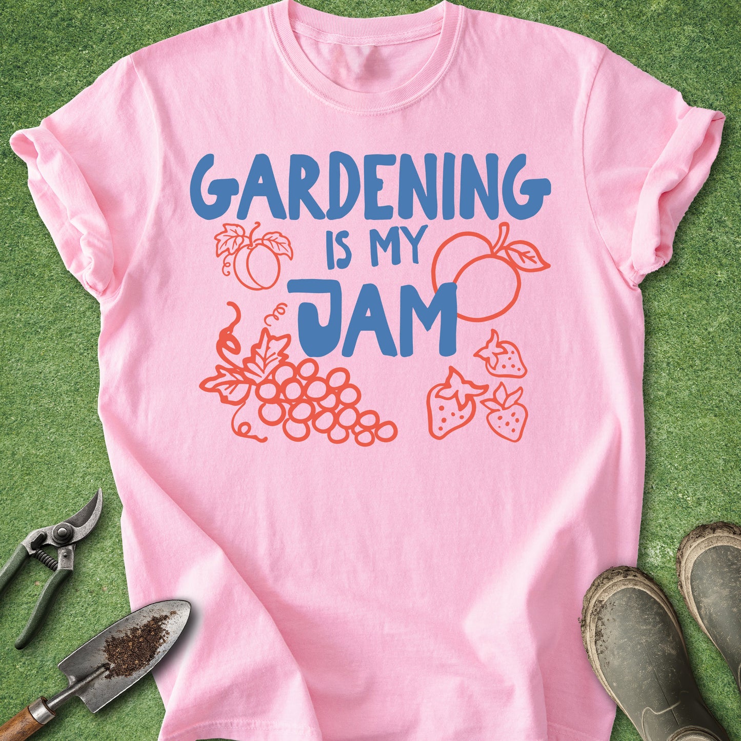 A pink short-sleeve T-shirt laid flat on green grass-like ground, with gardening tools and boots around it. The shirt has a large blue and orange graphic reading “GARDENING IS MY JAM” with fruit and plant illustrations.