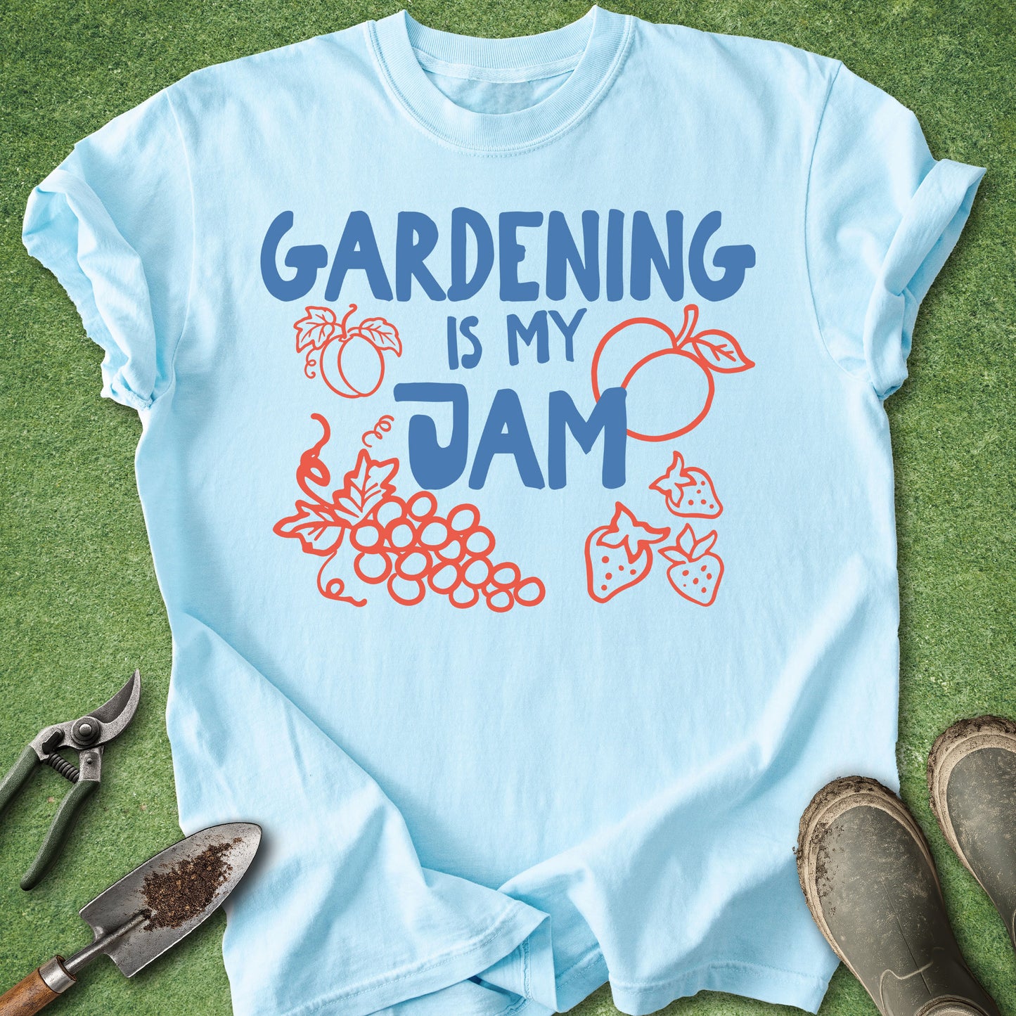 A light blue short-sleeve t-shirt laid flat on green grass, featuring the text 'GARDENING IS MY JAM' in blue with red/orange outline drawings of fruit and plants. Gardening tools and boots are placed around the shirt as styling props.