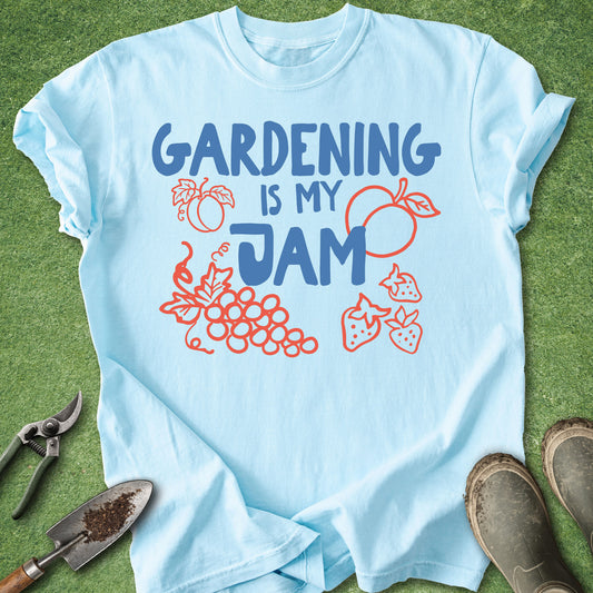 A light blue short-sleeve t-shirt laid flat on green grass, featuring the text 'GARDENING IS MY JAM' in blue with red/orange outline drawings of fruit and plants. Gardening tools and boots are placed around the shirt as styling props.
