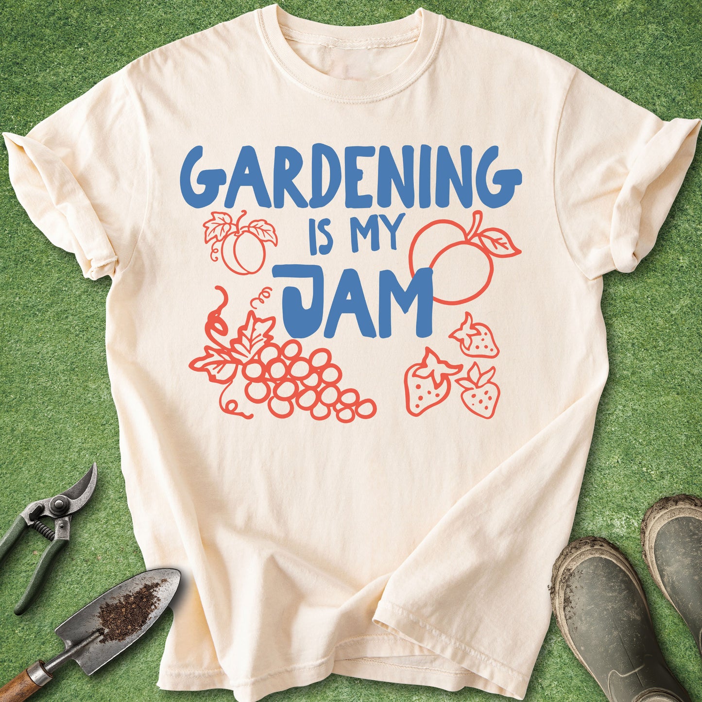 A cream-colored short-sleeve t-shirt with a graphic that reads 'GARDENING IS MY JAM' in blue, surrounded by red illustrations of fruits and vegetables, laid flat on green grass with gardening tools and boots nearby.