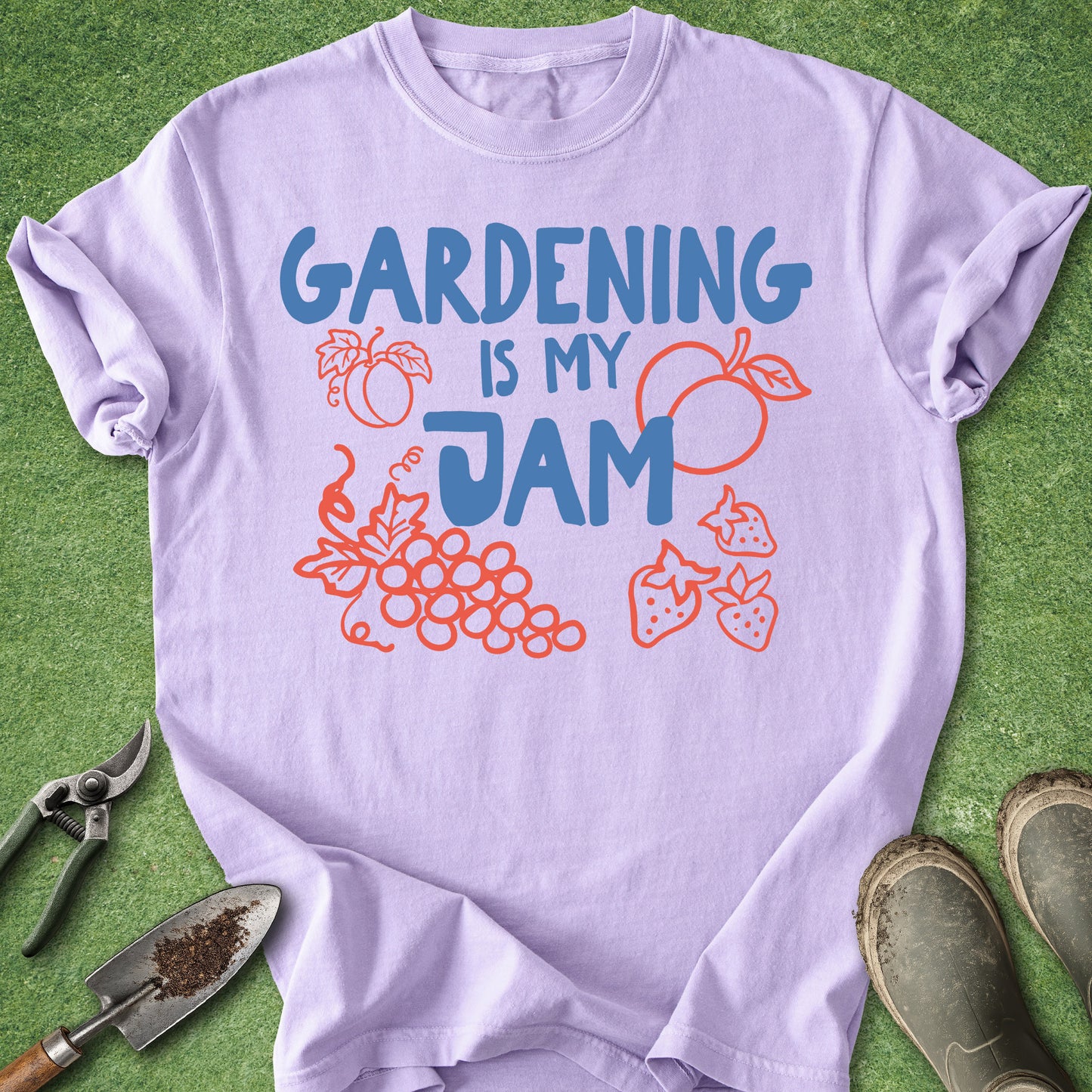 A light lavender t-shirt with a graphic that reads 'GARDENING IS MY JAM' in blue, styled in a flat-lay on green grass with gardening tools (trowel, pruning shears) and muddy boots.