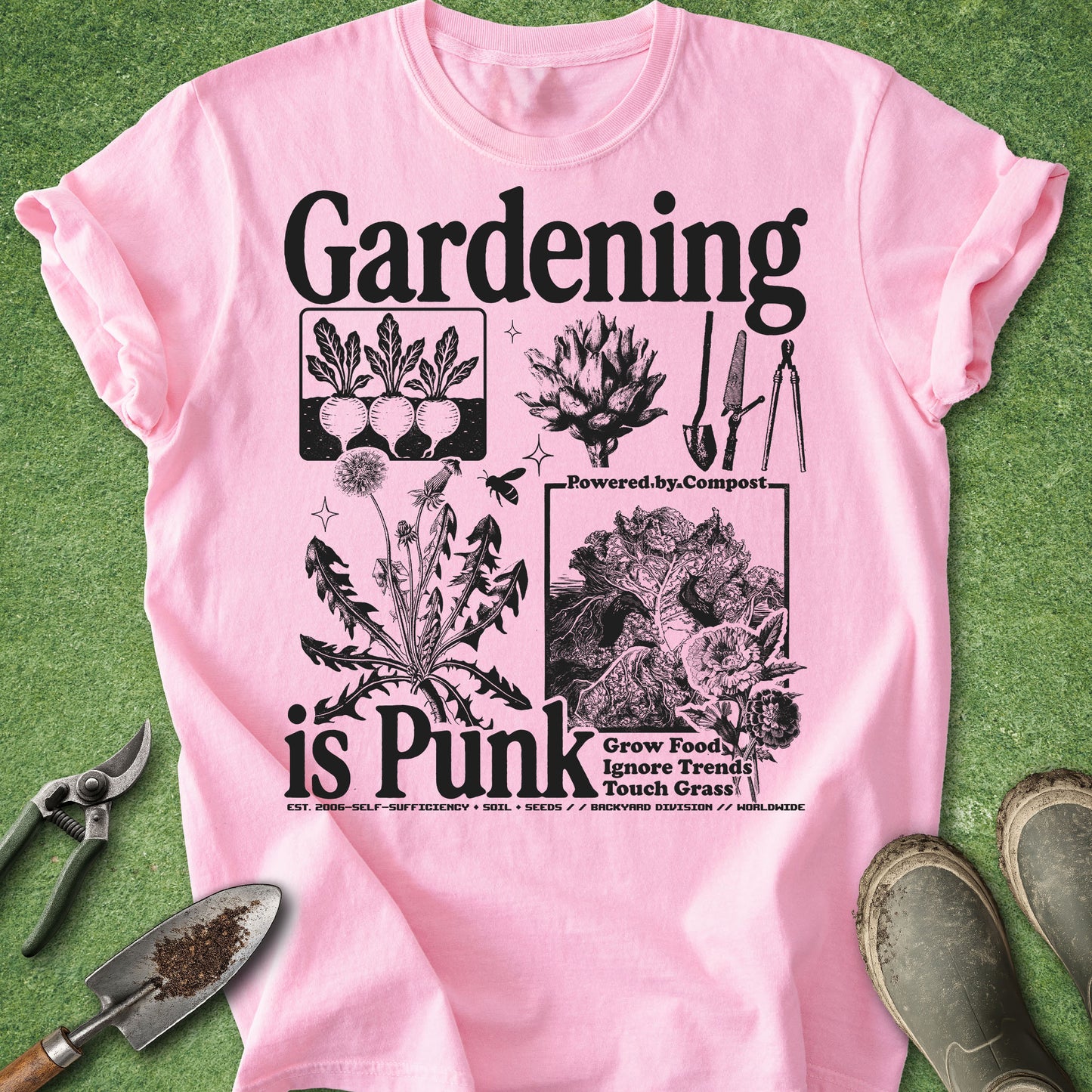 A light pink short-sleeve graphic T-shirt laid flat on a green grass-like surface, surrounded by gardening props including pruning shears, a trowel, and boots. The shirt has large black text reading 'Gardening is Punk' with illustrated plants, composting imagery, and smaller slogan text.