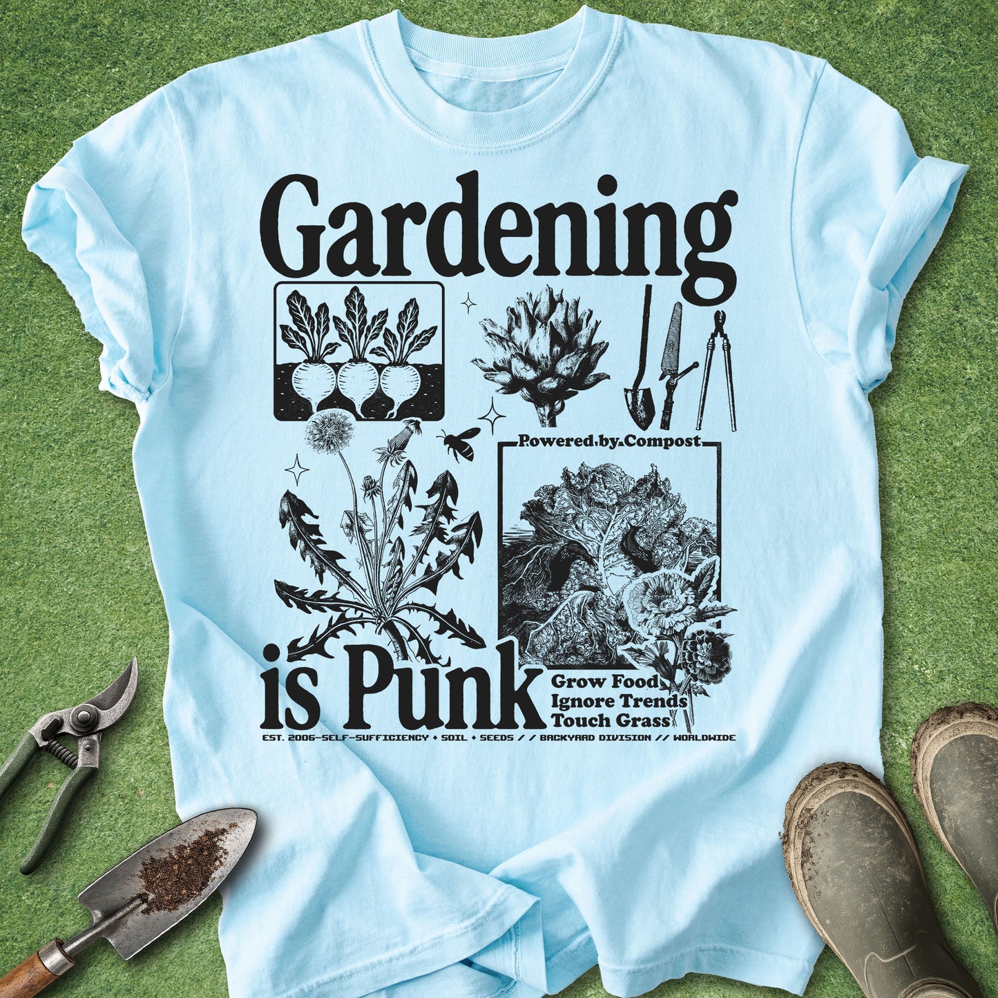 A light blue graphic t-shirt laid flat on green grass with gardening tools and boots around it. The shirt features a black graphic print that reads 'Gardening is Punk' with illustrations of plants, composting imagery, and gardening equipment.