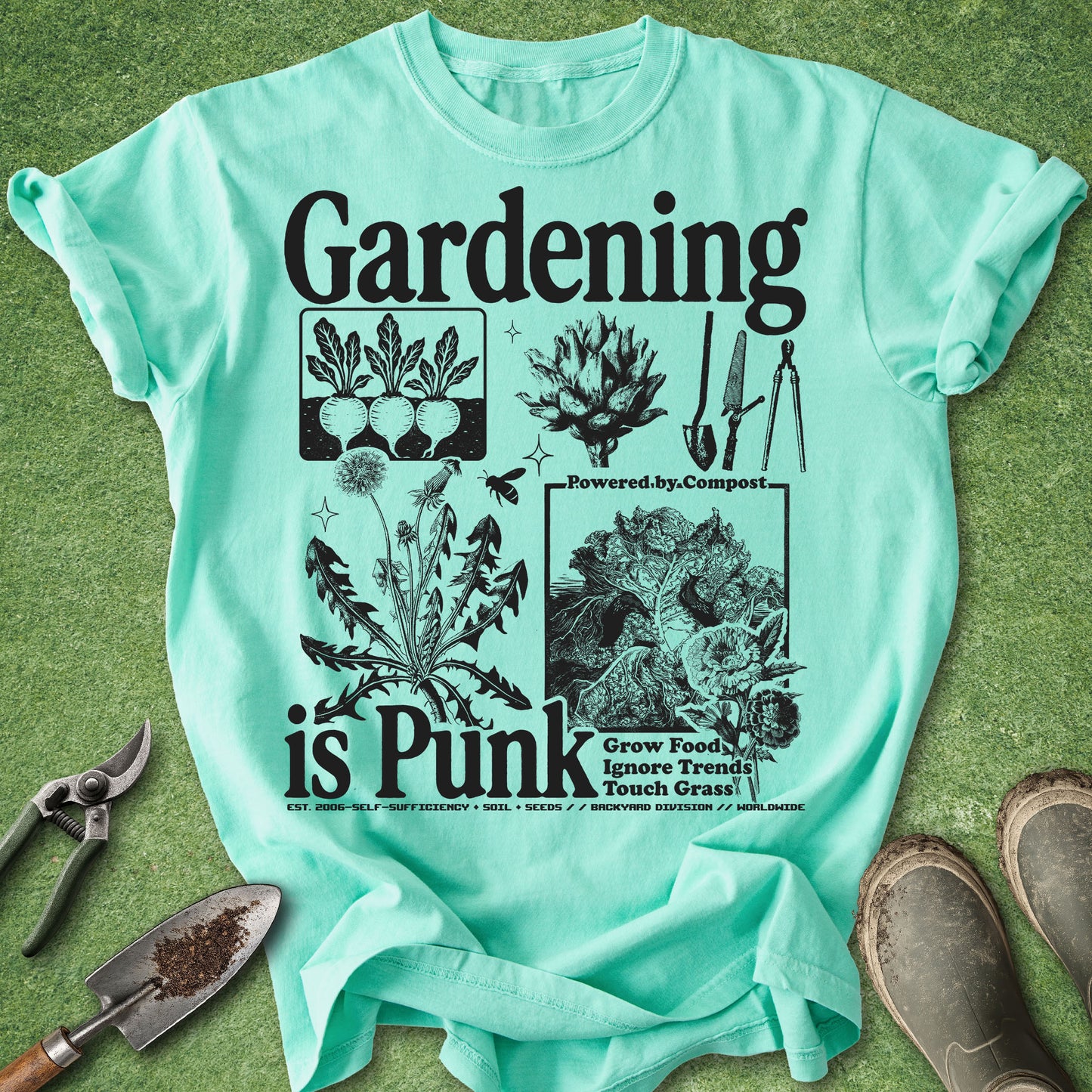 A mint green graphic t-shirt laid flat on a green grass-like surface, surrounded by gardening props including pruning shears, a trowel, and boots. The shirt features a black graphic print with the text 'Gardening is Punk' and illustrations of plants, composting, and gardening tools.