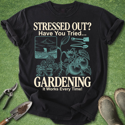 A black short-sleeve graphic T-shirt laid flat on green grass with gardening props around it. The shirt has a large cream and teal print reading “STRESSED OUT? Have You Tried.