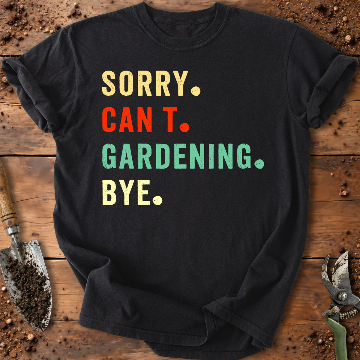 A black graphic t-shirt laid flat on a rustic wooden surface with soil and gardening tools. The shirt features the text 'SORRY.