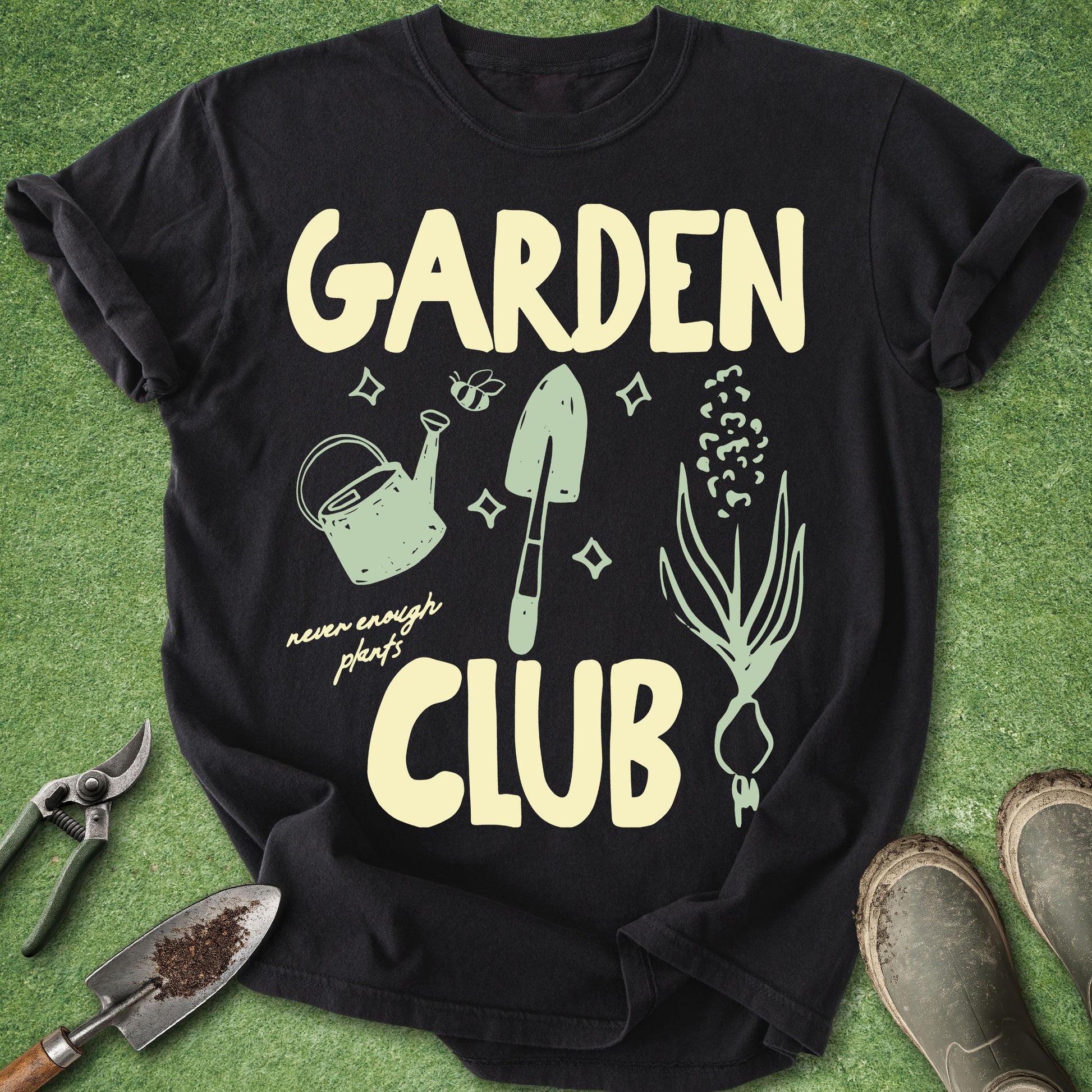 A black short-sleeve graphic T-shirt laid flat on green grass-like background, with large cream text reading 'GARDEN CLUB' and light green gardening illustrations including a watering can, trowel, seed, and onion. A pair of pruning shears and gardening boots are placed around the shirt as props.