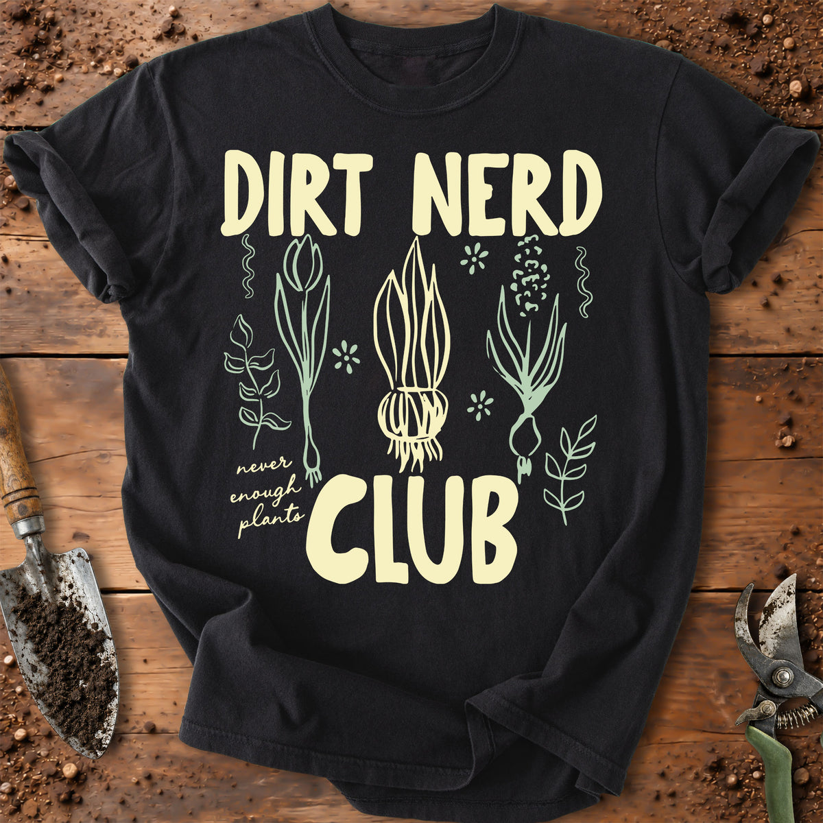 A black graphic t-shirt laid flat on a rustic wooden surface with gardening props like a trowel and pruning shears. The shirt features a cream-colored graphic with the text 'DIRT NERD CLUB' and illustrations of plants and an onion.