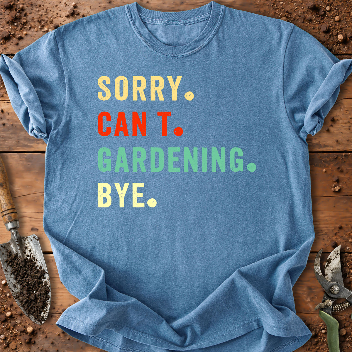 A light blue graphic t-shirt laid flat on a rustic wooden surface with gardening props like a trowel and pruning shears. The shirt features the text 'SORRY.
