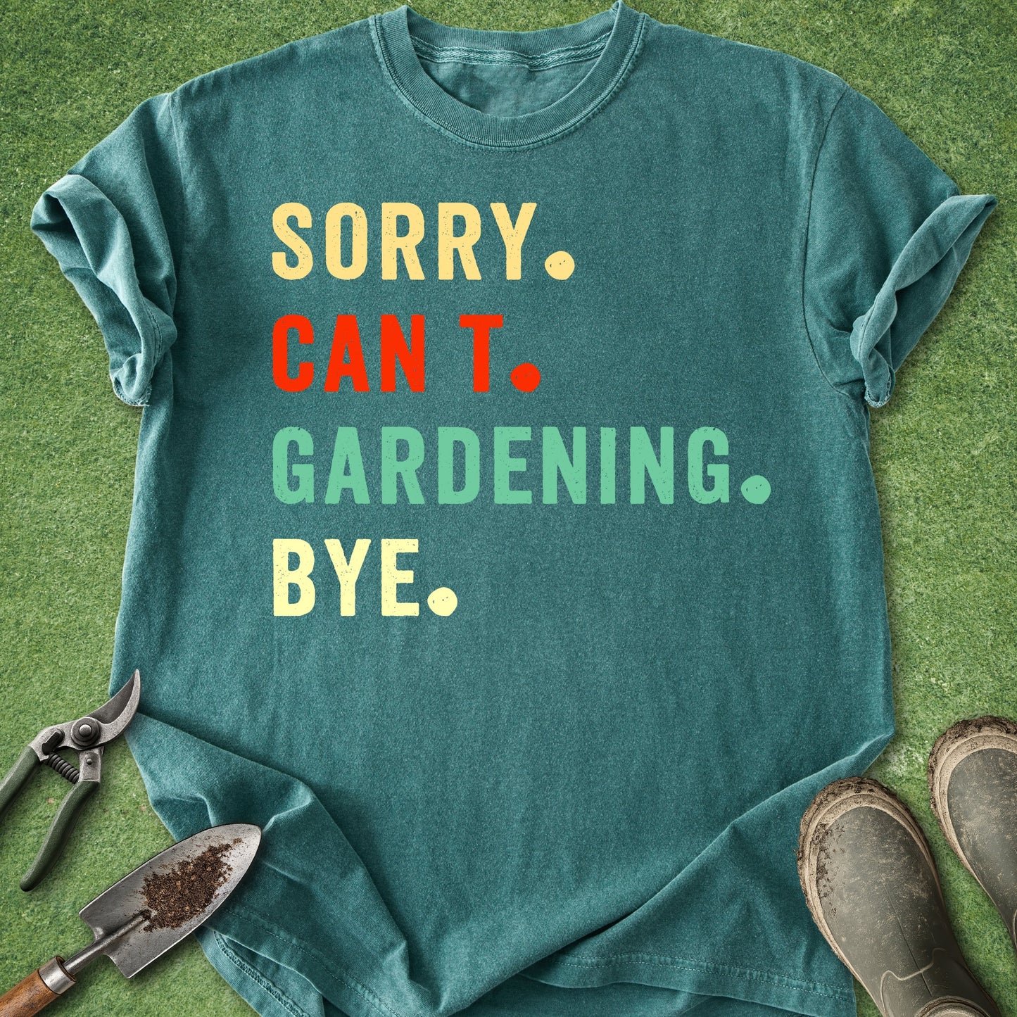 Teal t-shirt with colorful text on a grass background with gardening tools and shoes.