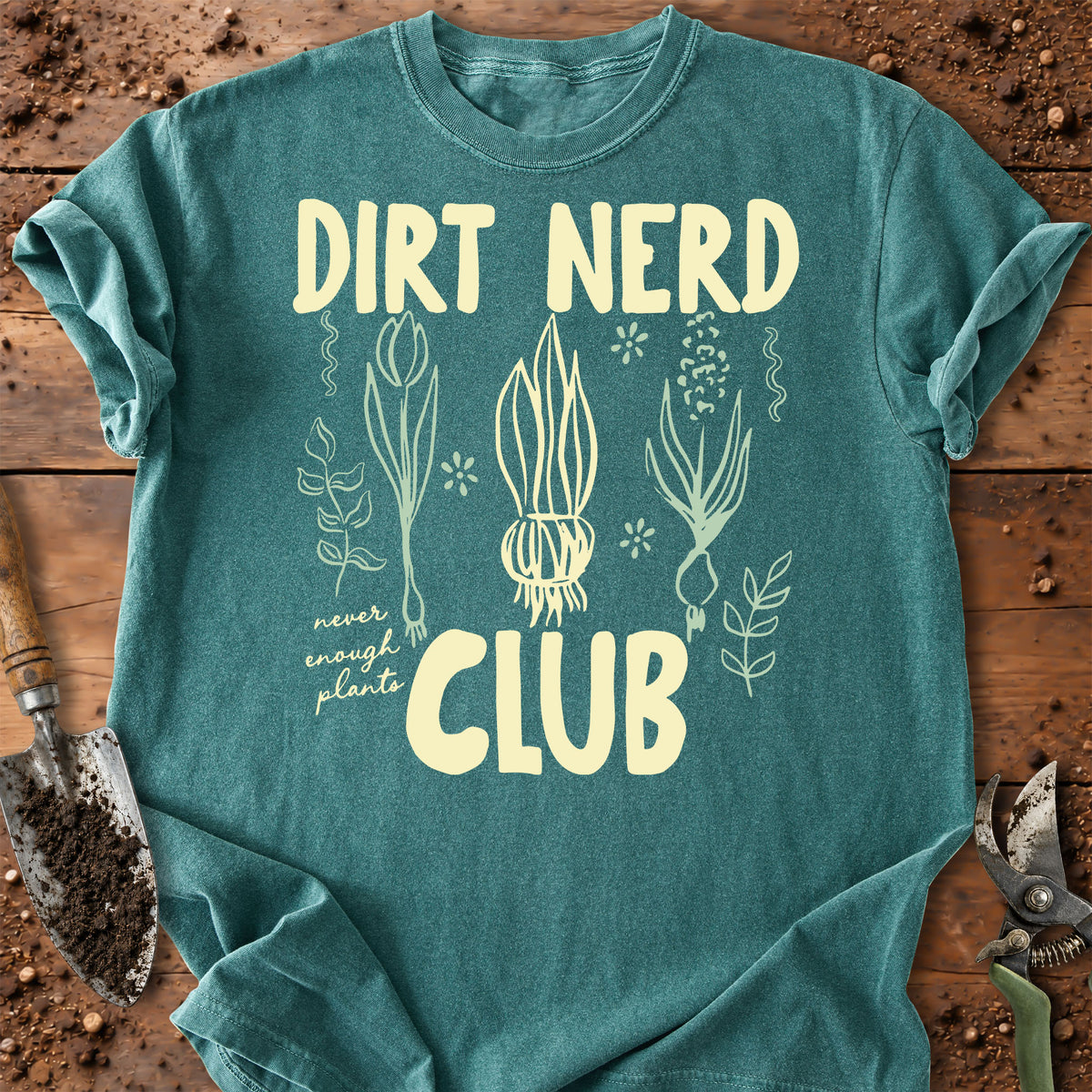 A teal t-shirt with a cream-colored graphic that reads 'DIRT NERD CLUB' surrounded by botanical illustrations, laid flat on a rustic wooden background with gardening tools.
