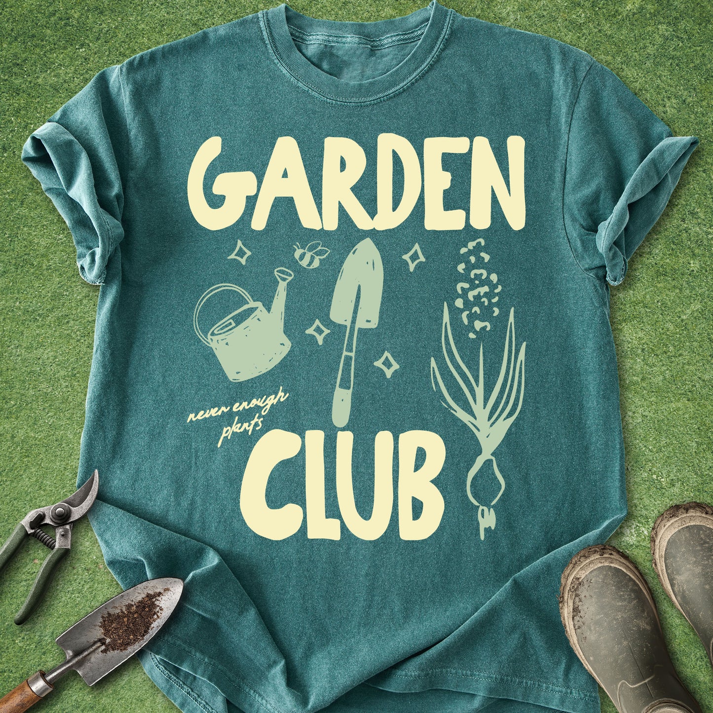 A teal short-sleeve t-shirt laid flat on green grass with gardening props around it, including pruning shears, a small trowel, and boots. The shirt has a large cream graphic reading “GARDEN CLUB” with illustrated gardening tools and plants and the phrase “never enough plants.
