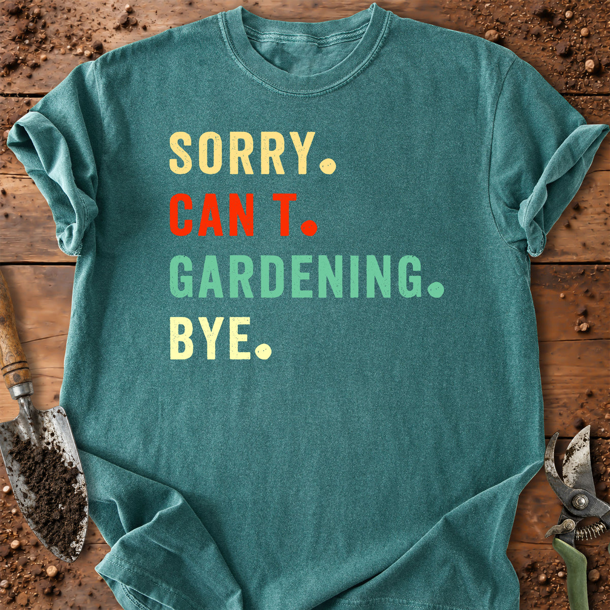 A teal graphic t-shirt laid flat on a rustic wooden surface with gardening props like a trowel and pruning shears. The shirt features the text 'SORRY.