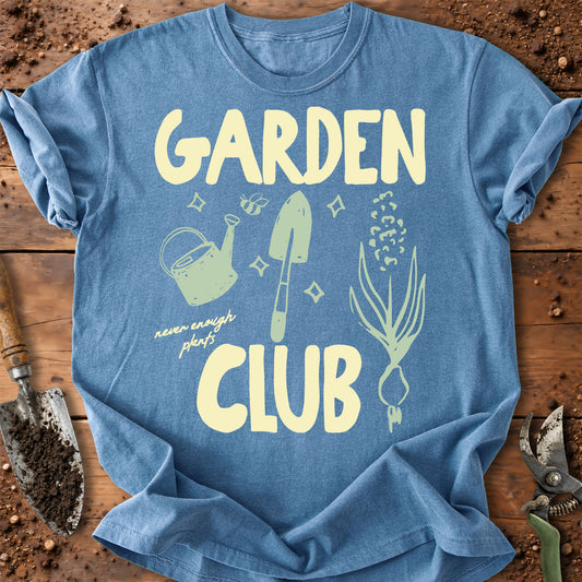 A light blue graphic t-shirt laid flat on a rustic wooden surface with gardening props. The shirt features the words 'GARDEN CLUB' in a pale yellow retro font, with a mint green watering can, shovel, plant sprout, and small text reading 'never enough plants'.