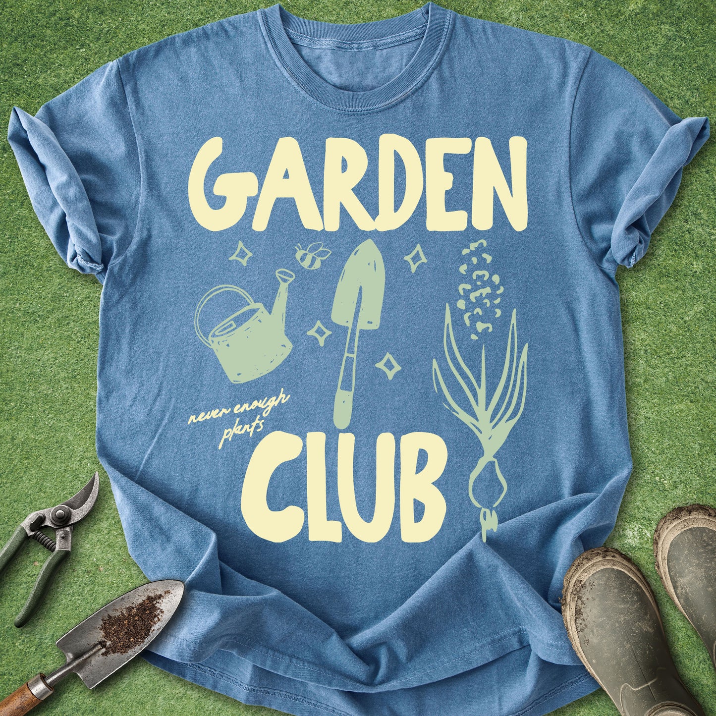 A blue short-sleeve graphic T-shirt laid flat on green grass-like ground, with gardening tools and boots around it. The shirt reads “GARDEN CLUB” in large pale yellow letters and includes a watering can, trowel, sprout, and small script text reading “never enough plants.