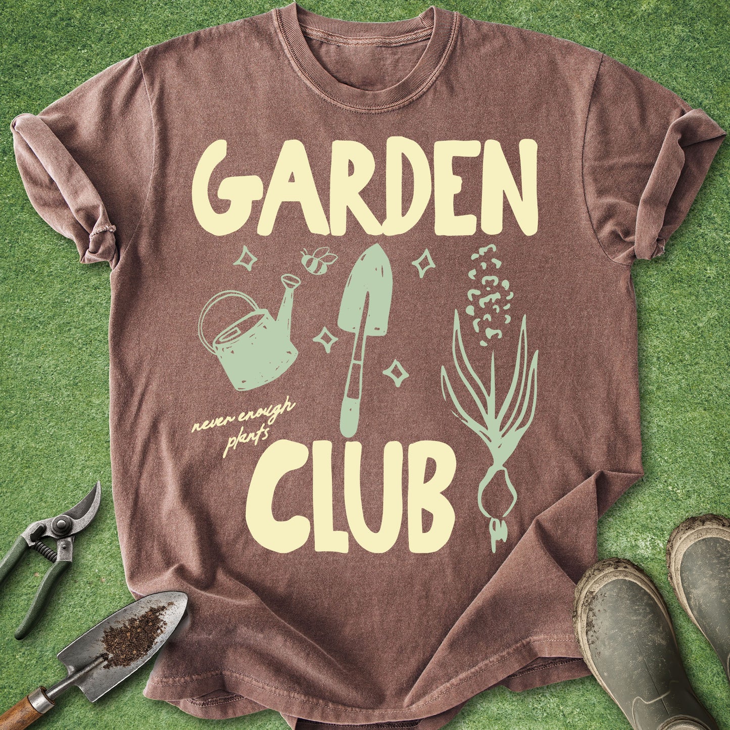 A brown short-sleeve t-shirt with a 'GARDEN CLUB' graphic print, laid flat on green artificial grass with gardening props like pruning shears, a trowel, and boots.